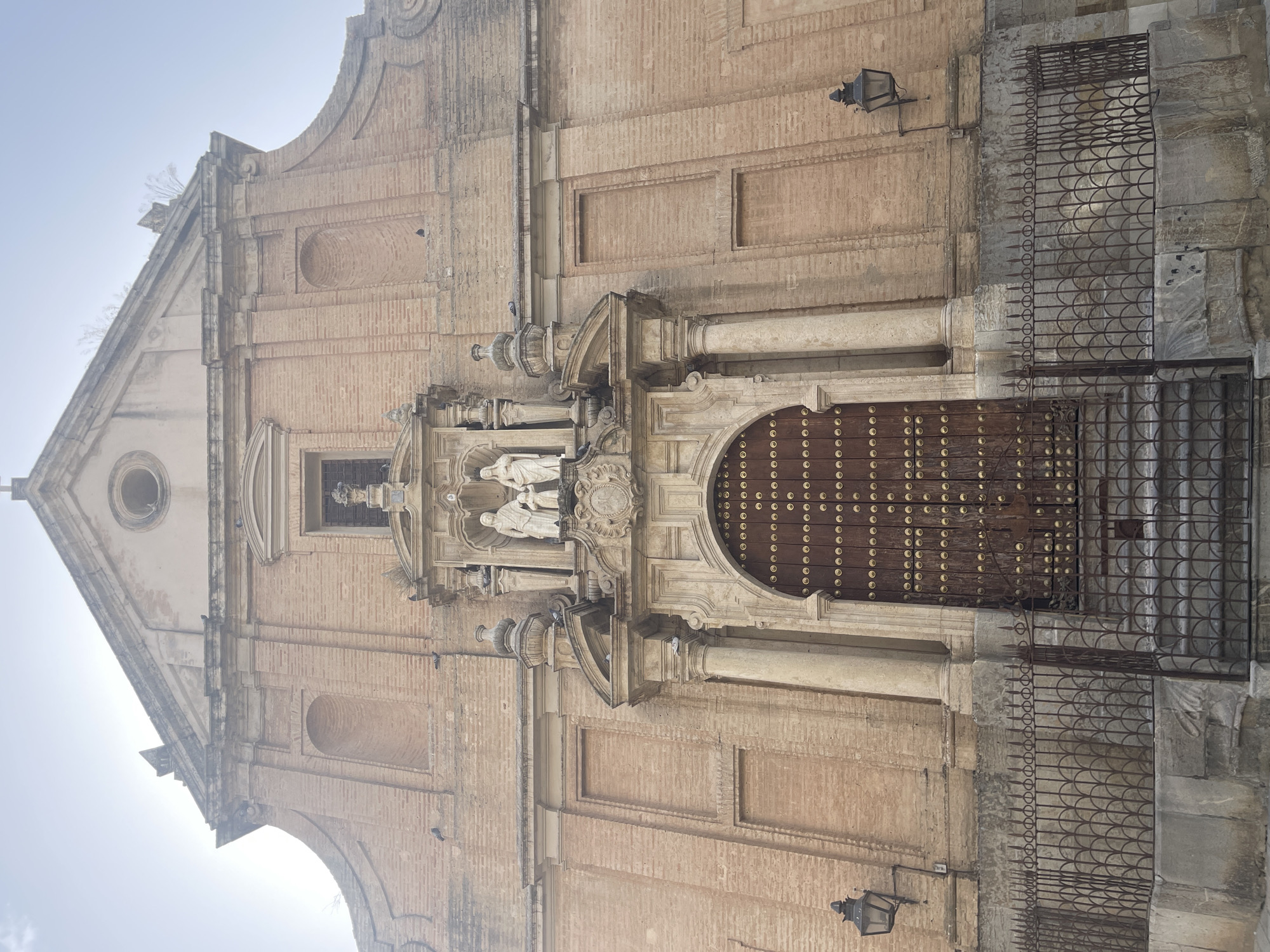 The image shows the facade of a brick church, likely in Córdoba, Spain, given the architectural style. The church has a large, ornate wooden door studded with metal embellishments, set within an arched entryway flanked by columns. Above the entrance, there are carved stone figures and decorative elements. The facade features a central pediment with a circular window and is constructed from reddish-brown bricks, with visible architectural details and some signs of weathering. Two black wrought-iron lamps are mounted on the exterior walls on either side of the entrance. A decorative wrought-iron fence runs along the base of the facade, separating it from a stone walkway with steps leading up to the entrance. Several pigeons are visible perched on the facade and ledges. The sky is overcast and bright.