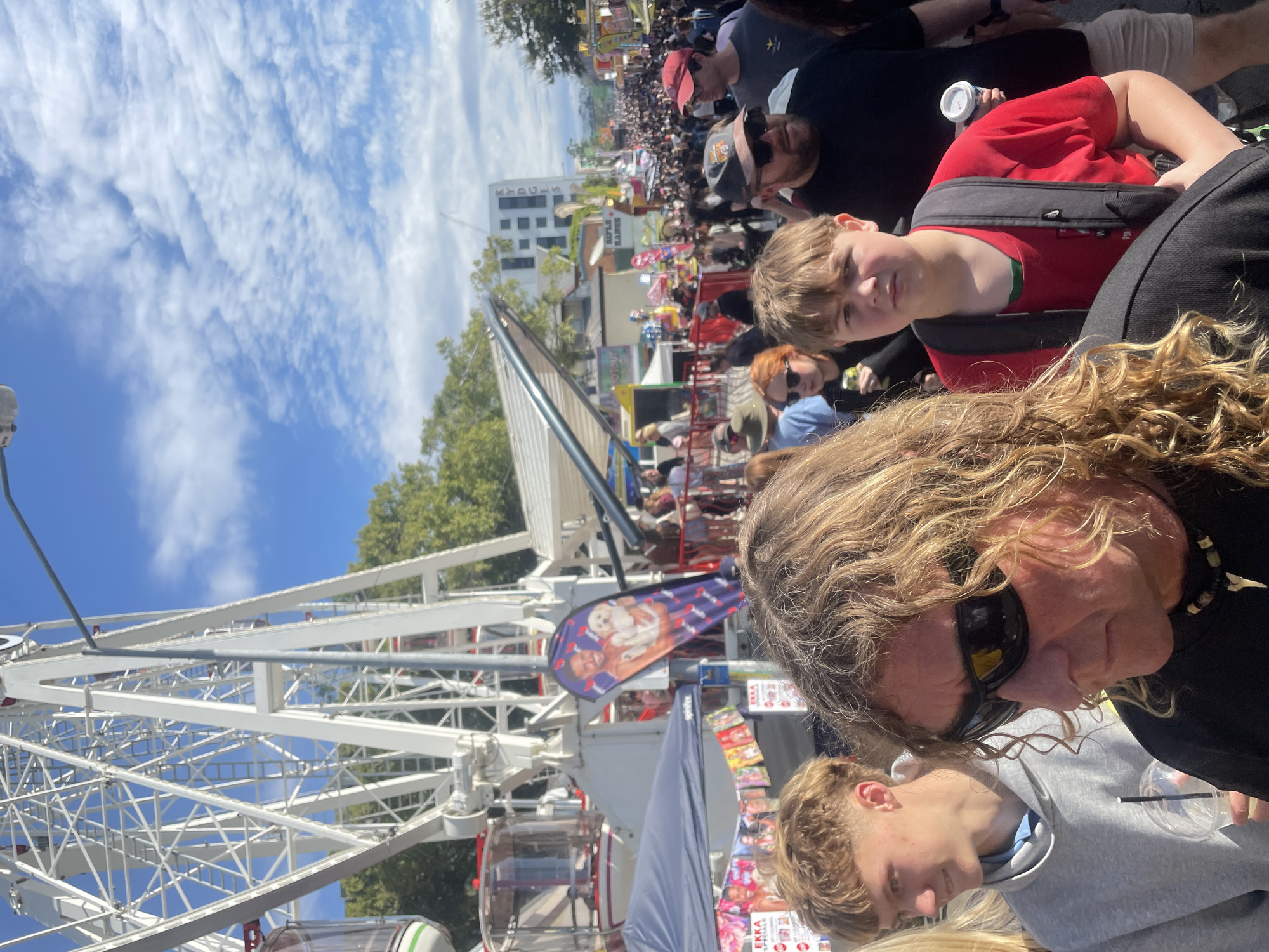 This image captures a crowded outdoor event, likely a fair or festival, with a large Ferris wheel prominently featured on the left. In the foreground, two young males are visible, with one partially obscured by the other. The central figure has light brown curly hair, is wearing dark sunglasses and a black t-shirt with a pendant necklace, and is holding a drink in a clear cup with a black straw. To his left, a younger male with short brown hair is wearing a grey hoodie and looking to his left.
In the background, the Ferris wheel's white structure dominates the scene, with some red and white cabins visible. A blue tent is set up near the base of the Ferris wheel, and a purple banner with an image of a man holding a dog is displayed. Further into the background, a throng of people walk along a street lined with various stalls and attractions. The street extends into the distance, creating a sense of a bustling environment. Buildings are visible in the far background, with one prominent white building featuring the word "RYDGES" in vertical lettering, suggesting this location is in Brisbane, Australia, specifically near the Rydges South Bank hotel. The sky is blue with scattered white clouds, indicating it is daytime. The overall impression is a lively public gathering on a sunny day.