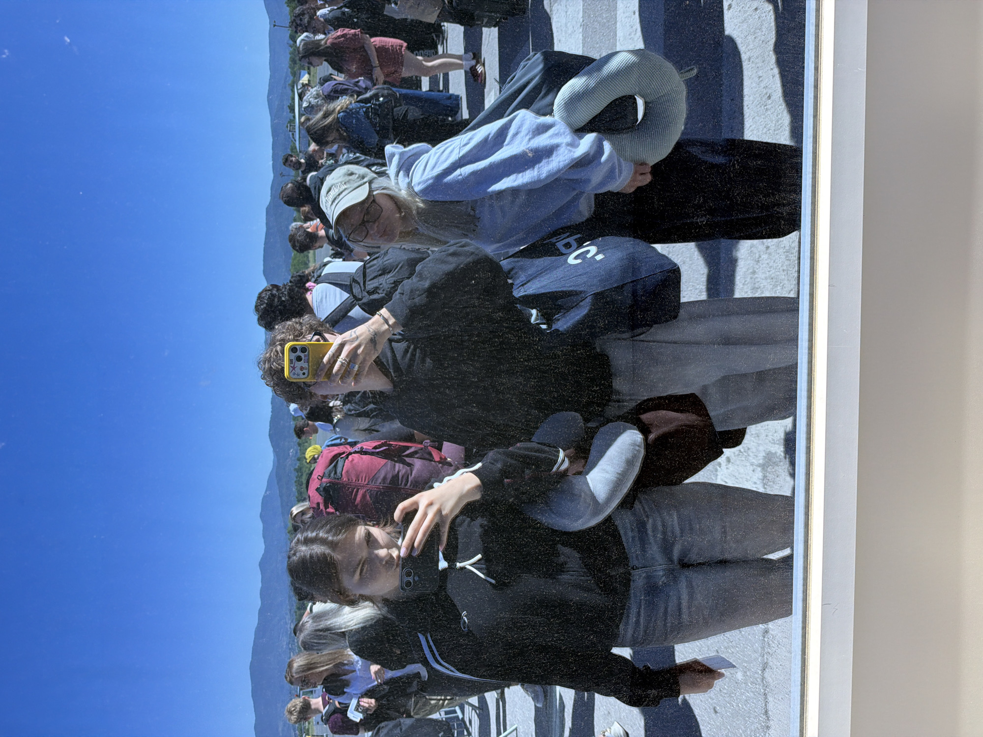 A photograph depicts a group of people, reflected in glass, with a backdrop of a clear blue sky and distant mountains. The reflection suggests the individuals are in an outdoor environment. One person in the foreground is holding a yellow mobile phone, presumably taking a picture. Another individual is wearing a black jacket and jeans, cradling a gray travel pillow. A third person wears a light blue sweatshirt and black pants, also holding a light-colored travel pillow. The presence of travel pillows and luggage indicates the group may be in transit, possibly at an airport or travel hub. The city of Gornje Dubrave, Bosnia and Herzegovina, is visible in the reflection. The reflection also shows the edge of the glass surface and a white wall.