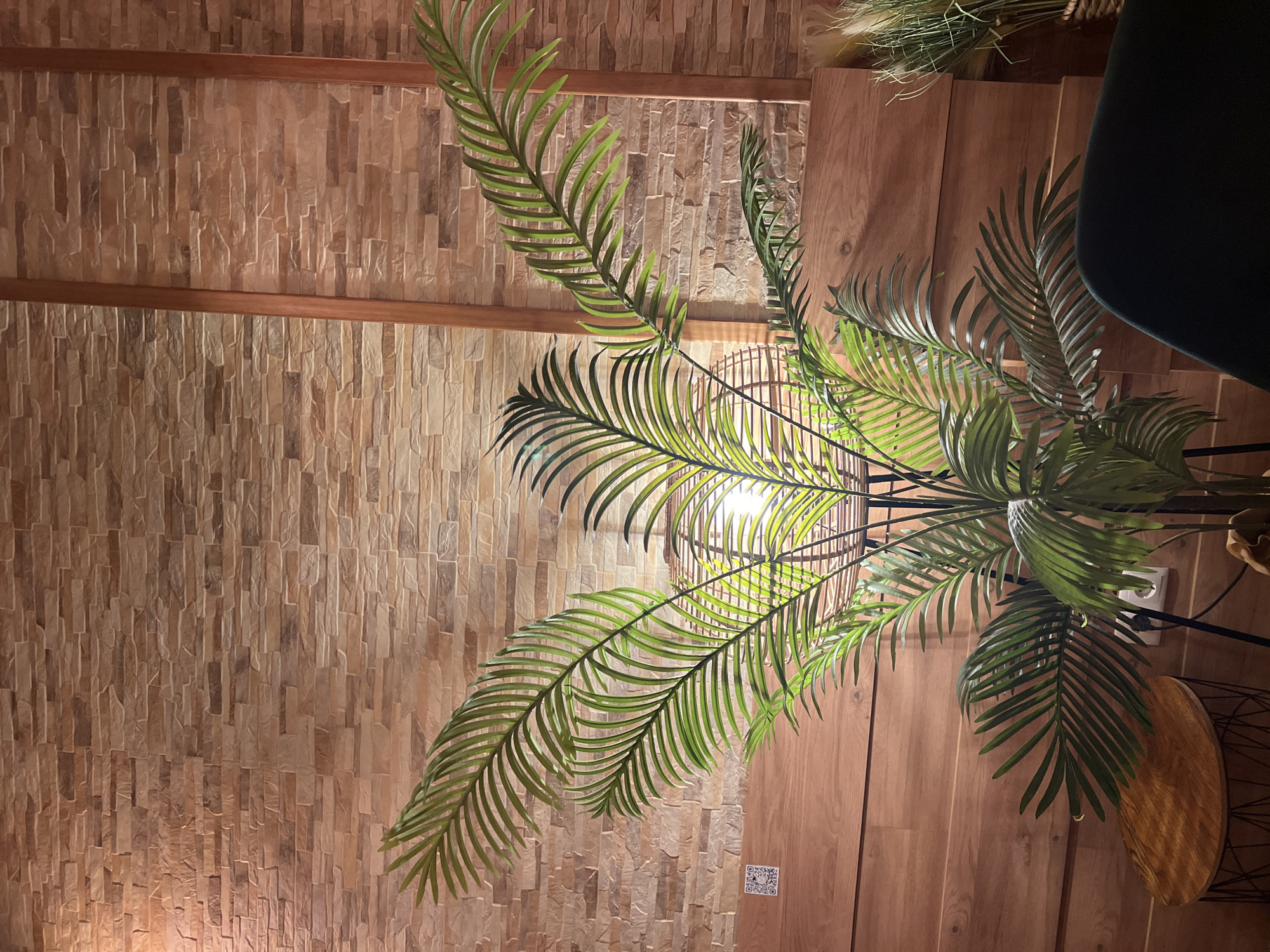 The image captures a low-angle shot, likely from within a building, focusing on interior design elements. The primary subject is a decorative palm plant with vibrant green fronds, centrally positioned in the frame. A cylindrical light fixture with a dark, open-weave frame is partially visible behind the plant.
The background consists of two distinct wall sections. To the left, a textured, beige brick-patterned wall extends vertically. Above it, a wooden beam spans horizontally. To the right, a smoother, warm-toned wooden wall is present.
A small, almost square QR code is seen on the right-hand wooden wall. There is also a partially visible electrical outlet near the base of the plant. A dark green chair or piece of furniture is partially visible at the top right corner. At the top, some grassy decorations are also seen.
The image includes a warm color palette, indicating interior lighting is likely artificial.
The location is in Sant Pere I Sant Pau, Spain.