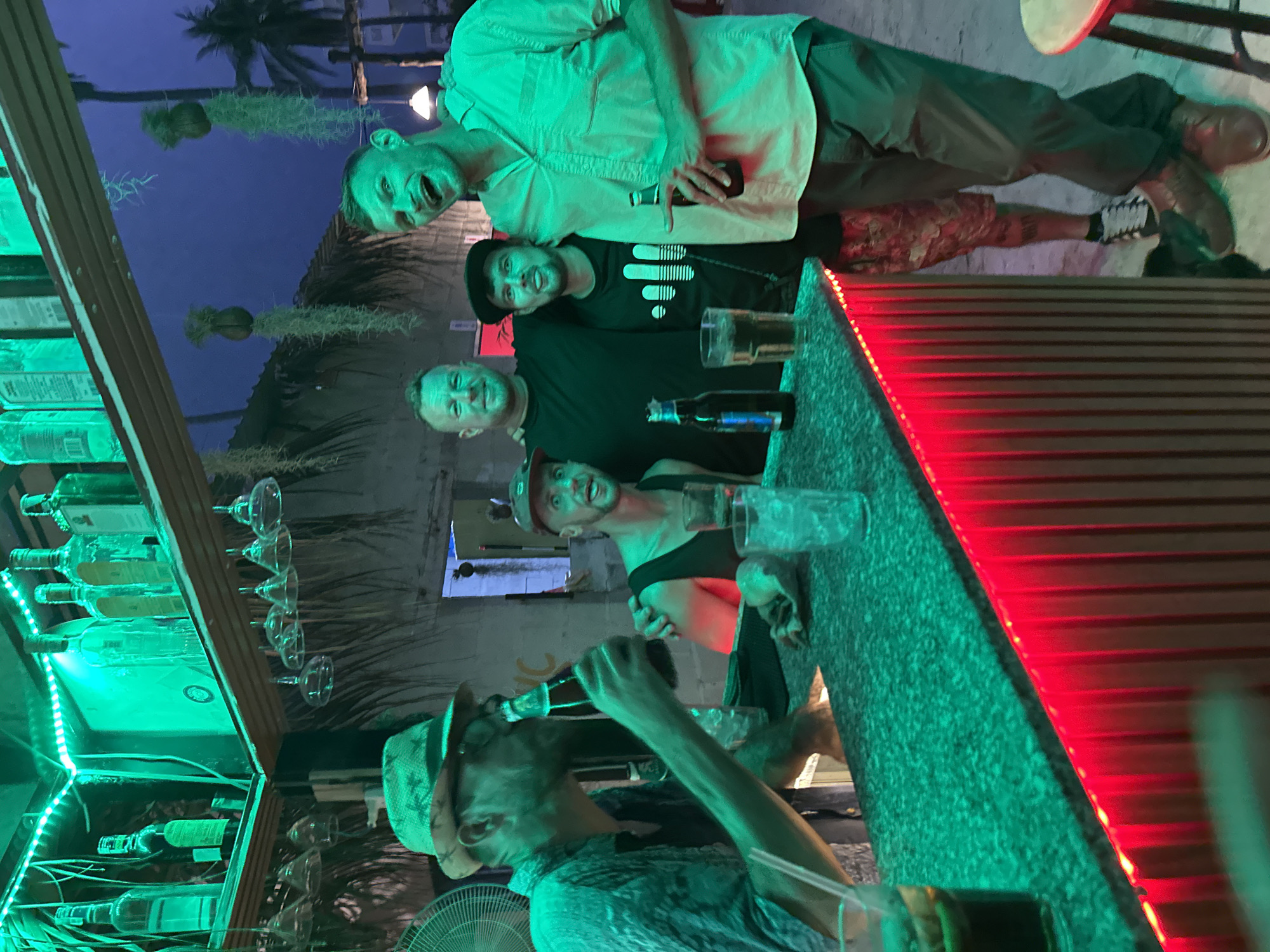 Five men are standing at a bar in Mũi Né, Vietnam. The bar has a green-lighted shelf of liquor bottles and glassware on the left and a red-lighted counter on the right. The men are posing for the photo, with the front one drinking from a bottle. The background shows a thatched roof building and palm trees. The lighting in the bar creates a festive atmosphere.