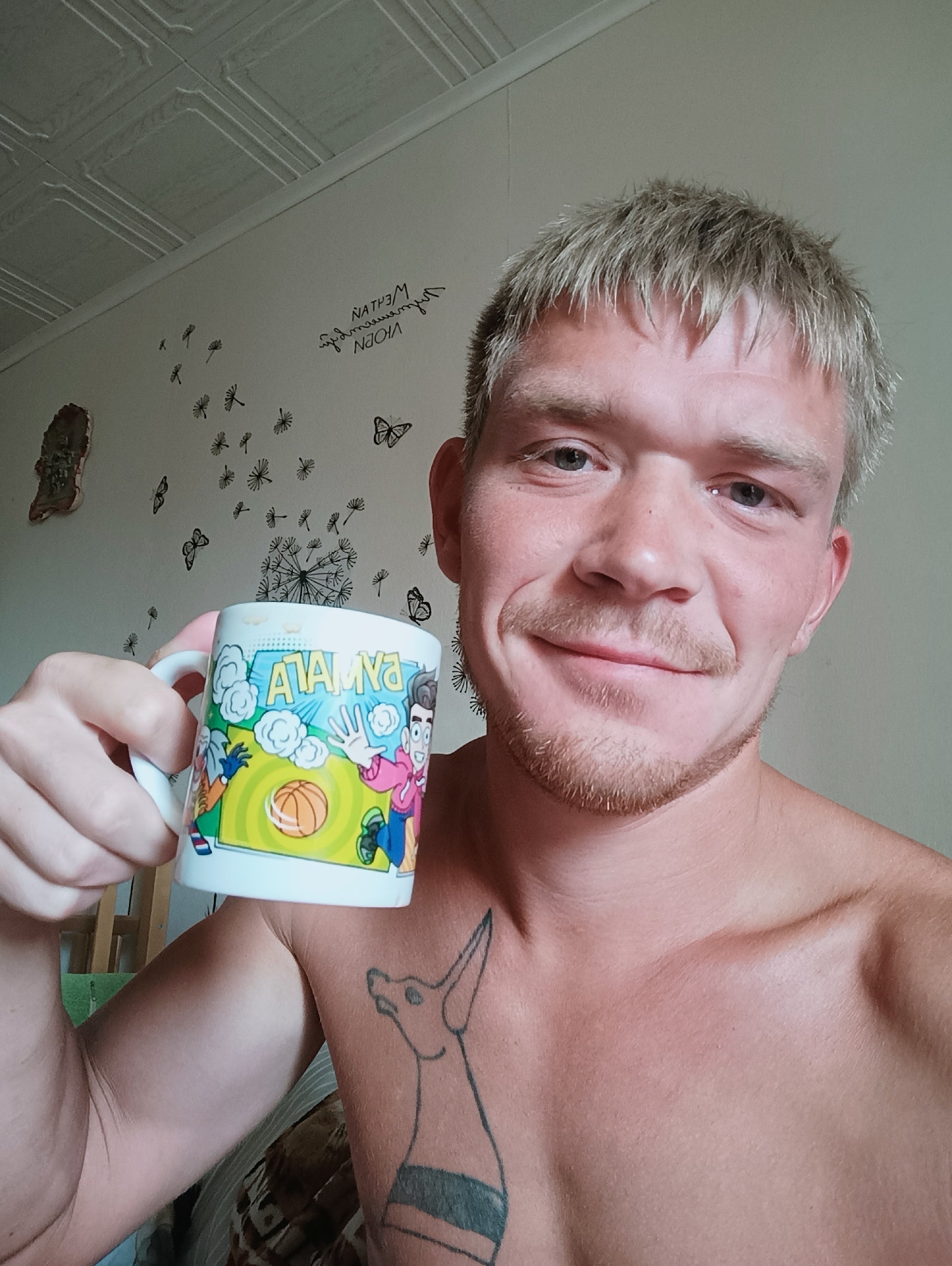 A man with short, blonde hair and a light beard holds a white mug with a cartoon drawing of basketball players and the word "ATAMNA" on it. He is shirtless, revealing a tattoo of a dog on his chest. The background consists of a light-colored wall with wall decals of butterflies and dandelions, and a textured ceiling. The setting appears to be indoors, possibly a residential room, with a wooden chair or crib visible in the background on the left. The image was taken in Chaikovsky, Russia.