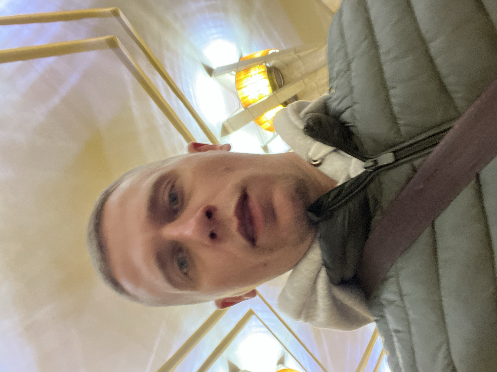 A male individual with short, light-colored hair and blue eyes is depicted in a close-up, low-angle selfie. He is wearing a grey hooded sweatshirt layered under an olive-green puffer jacket, with a dark strap visible across his upper chest. The subject maintains direct eye contact with the camera. The background reveals the ornate interior of Ploshchad Yakuba Kolasa Metro Station in Minsk, Belarus. Distinct architectural features include a light-colored ceiling adorned with geometric gold-toned linear patterns and multiple large, stylized light fixtures. These fixtures emit a warm, diffused yellow light, illuminating the cream and beige overhead structures.