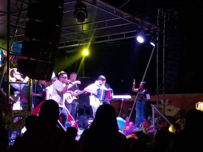 A band performs on an outdoor stage lit with spotlights, featuring a lead singer with a microphone, a guitarist, and an accordion player. A woman stands to the side, holding a cell phone up as if recording or taking a picture. A crowd of people is visible in the foreground, partially obscuring the view of the stage. Brightly colored balloons are scattered among the audience. Stage equipment and scaffolding are visible above the band.