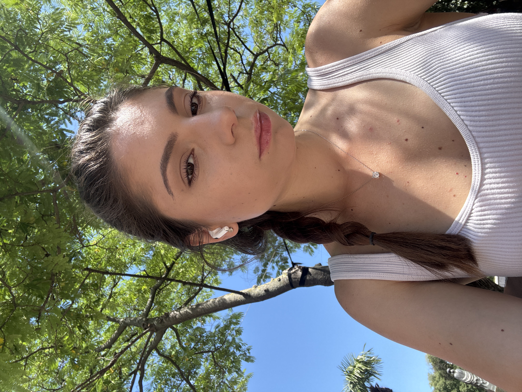 A young woman with brown hair in a braid is looking towards the camera, her face slightly turned to the side. She is wearing a white ribbed tank top and a silver necklace with a pendant. She also has white earbuds in her ears. The background shows a tree with green leaves and a bright blue sky. The location is the Sea Garden in Varna, Bulgaria.