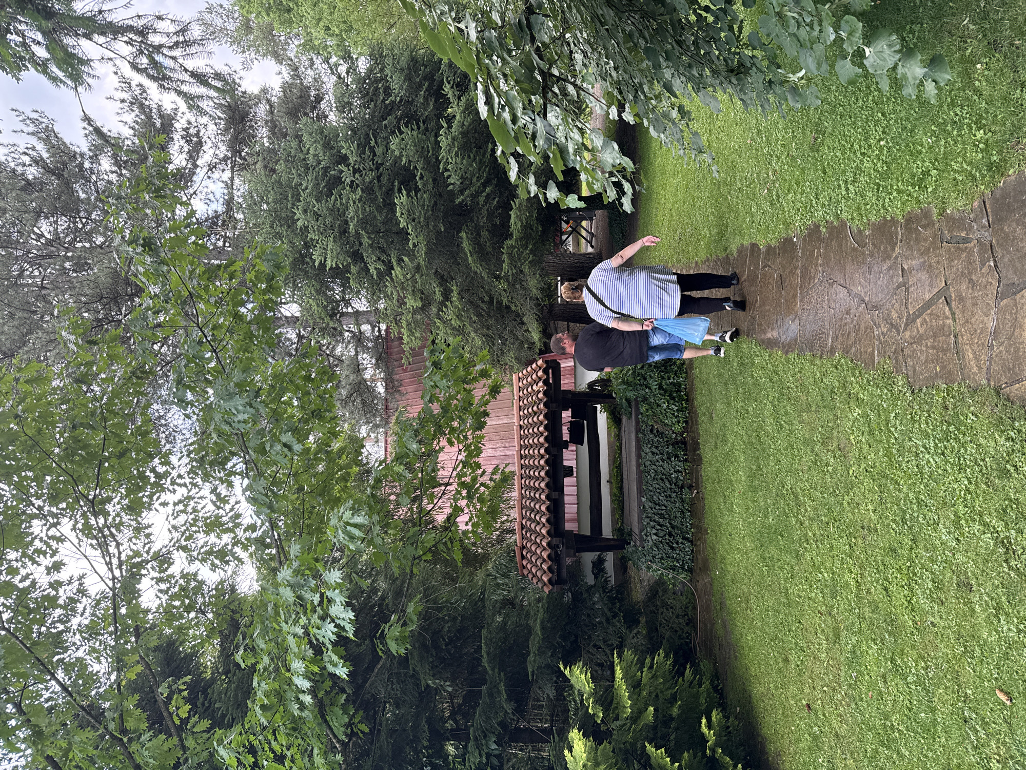 The image depicts an outdoor scene in Tarnicheni, Bulgaria, featuring a lush, verdant environment. A wet, irregular flagstone path extends from the foreground into the midground, gently curving to the right. Two individuals are walking away from the viewer on this path. The person on the left, a male, wears a black t-shirt, blue denim shorts, and dark athletic shoes, carrying a light blue plastic bag in their left hand. The person on the right, a female, wears a white and blue striped short-sleeved top, dark pants, and dark flat shoes.
The path is flanked by well-maintained green grass. To the left of the path, in the midground, a rustic well-like structure is present, featuring a terracotta-tiled roof supported by wooden beams. A dark, bell-shaped object hangs from one of the beams. Behind this structure, a low wall or base is covered with ivy.
The area is densely vegetated with a variety of trees and shrubs. Large deciduous trees with green leaves, possibly oak, are prominent in the upper left and foreground, framing the scene. Coniferous trees, including firs and pines, are scattered throughout the midground and background, with some tall specimens reaching high into the cloudy sky. A building with a reddish-orange corrugated metal roof and lighter walls is partially visible in the background, mostly obscured by the tree canopy. The wetness of the path and the overcast sky suggest recent rain or damp conditions.