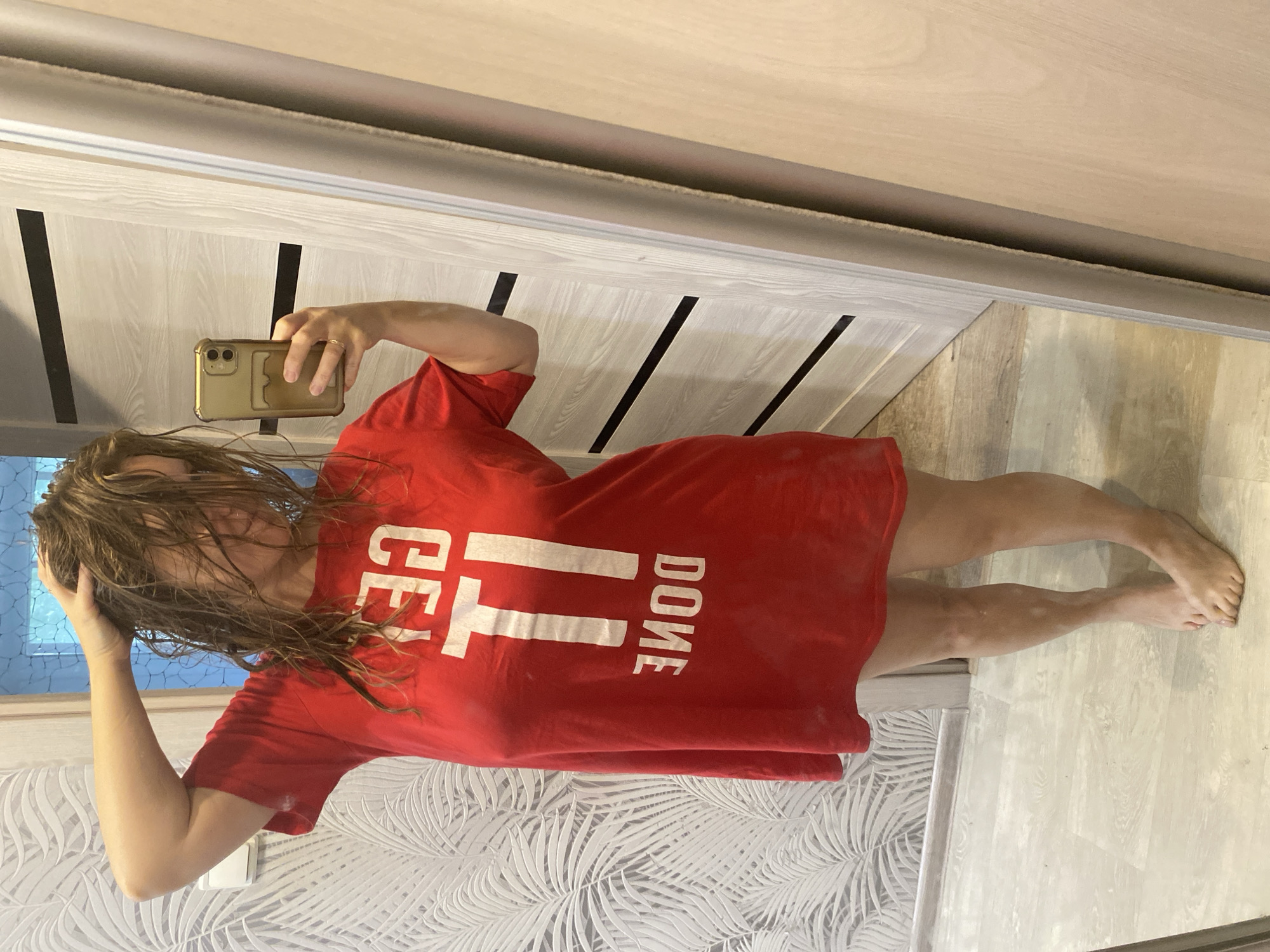 A woman stands in what appears to be a residential interior, taking a selfie in a mirror. She is wearing a red t-shirt with white lettering that reads "GET IT DONE". Her hair is wet and she is holding her head with one hand. Her legs and feet are visible, and she is barefoot. The background includes a wall with a palm leaf pattern wallpaper, a light-colored door with horizontal black stripes, and a large mirror reflecting the scene. The floor is a light-colored laminate. There are no visible indications that this location is in Noviy, Russia.