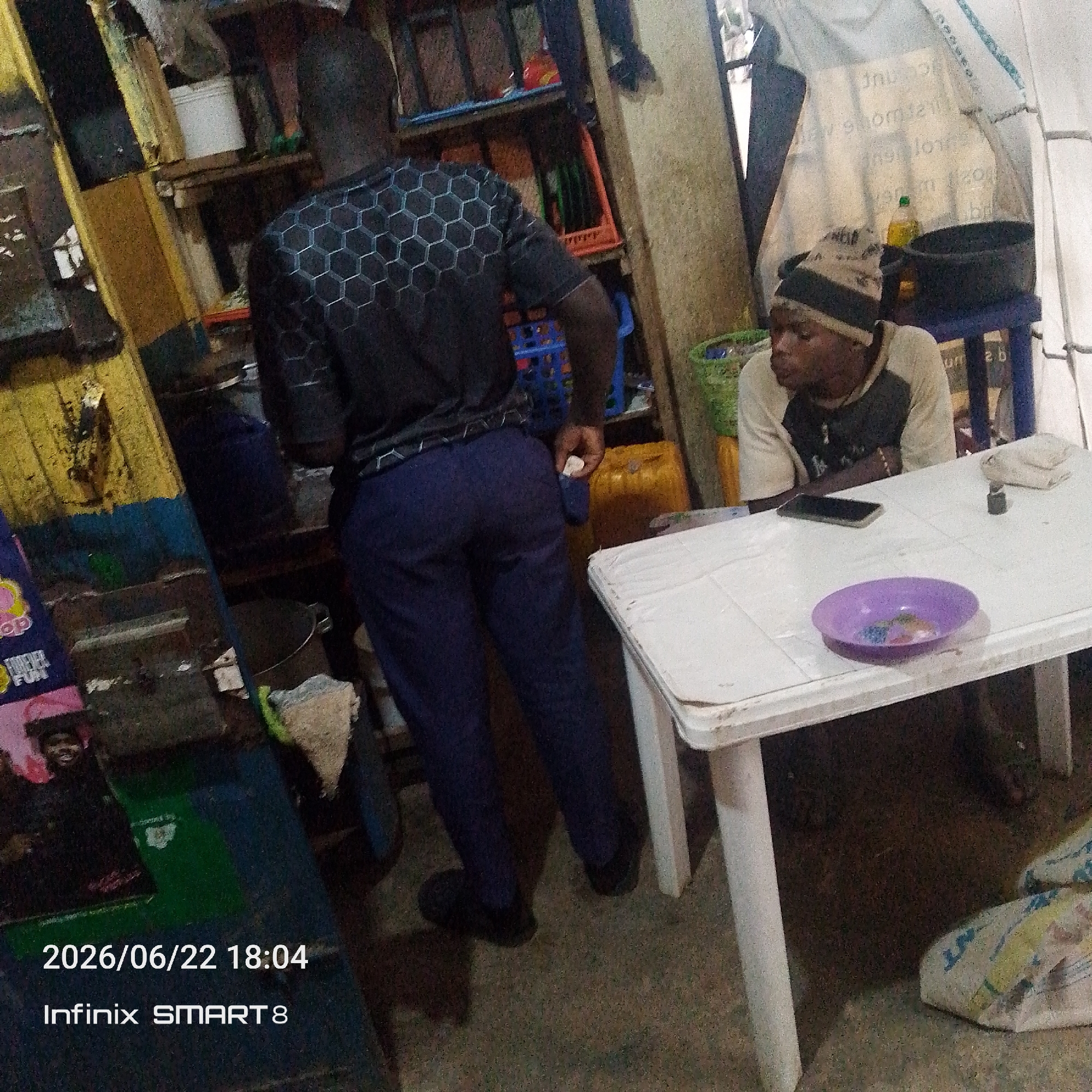 In an indoor setting with cluttered shelves and a general air of disarray, a person stands facing away from the camera, wearing a dark shirt with a hexagonal pattern and dark blue pants. To their right, another person wearing a beanie hat and a light-colored shirt is seated at a white plastic table. On the table, a purple bowl containing small colored objects, a smartphone, and a small metal object are visible. The environment appears to be a market or stall area, with various containers and items present on shelves and on the ground. The timestamp "2026/06/22 18:04" and "Infinix SMART 8" are overlaid on the bottom left of the image.