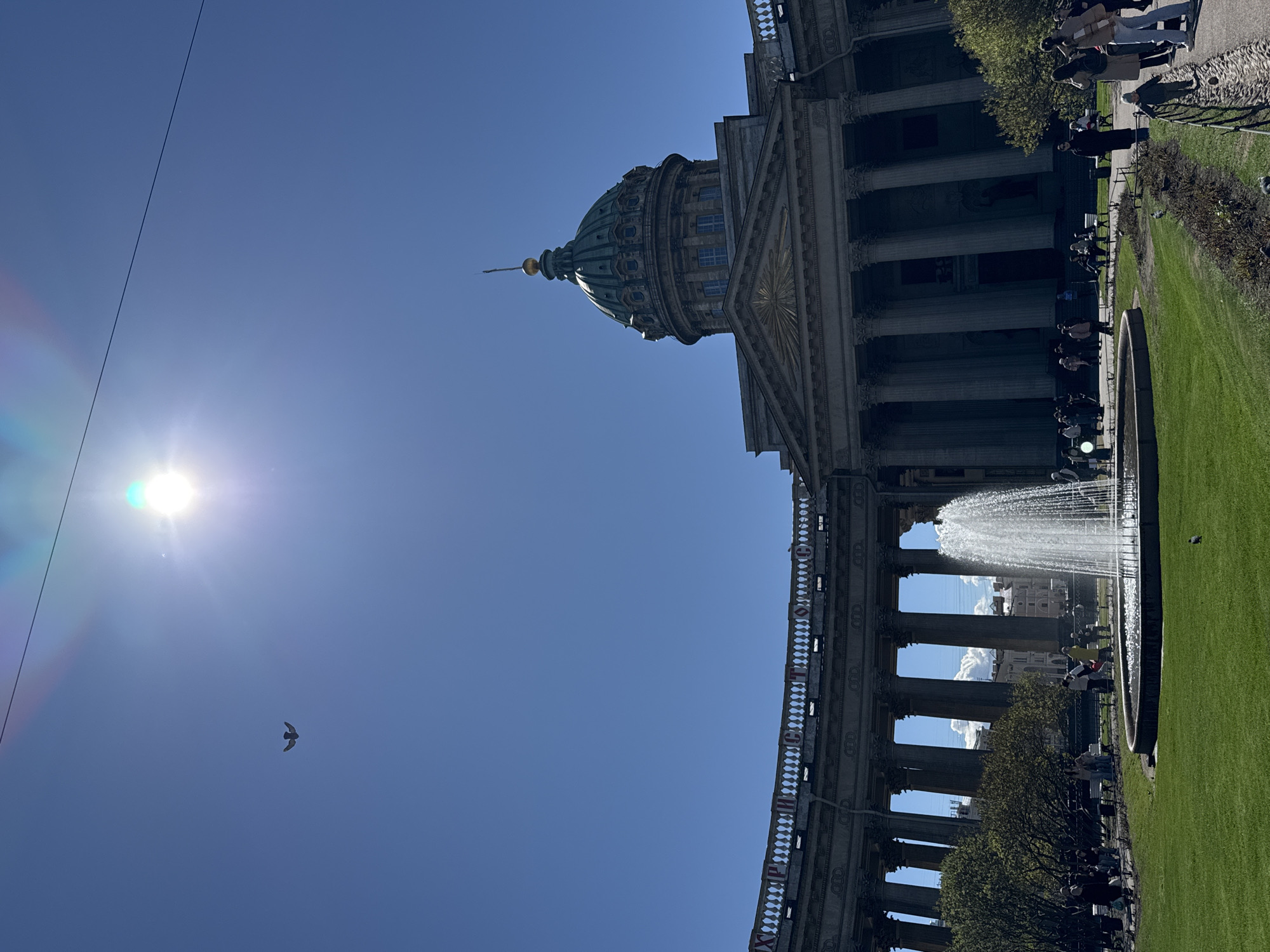 The image depicts a low-angle view of the Kazan Cathedral in Saint Petersburg, Russia, under a bright, clear sky with the sun visible. The cathedral's dome and colonnade are prominent, as is a fountain situated in front of the building. People can be seen strolling and sitting near the fountain and along the lawn in front of the colonnade. A bird is flying in the sky between the sun and the dome.
