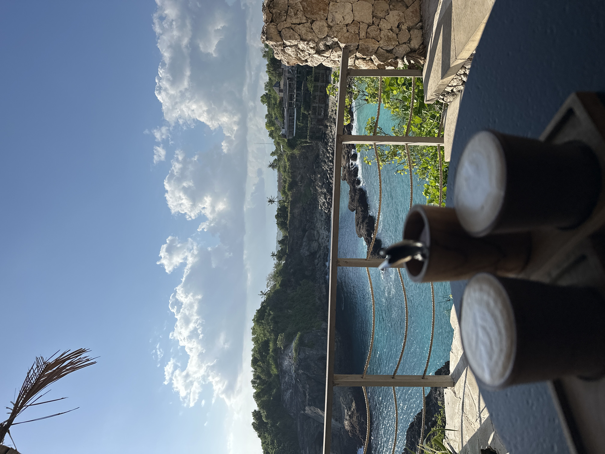 The image depicts a scenic view of a rocky coastline with turquoise water under a bright blue sky with scattered white clouds. In the foreground, a dark blue surface, likely a table, is visible, with three cylindrical objects made of wood or bamboo resting on it. One of these objects has a circular pattern on its surface. A railing with a rope detail separates the foreground from the view of the coastline.
The coastline is covered with lush green vegetation and features steep cliffs. Some structures, possibly buildings or parts of a resort, are nestled into the hillside. The sea is a vibrant blue, with visible waves crashing against the rocks at the base of the cliffs. The overall setting suggests a tropical or semi-tropical location, reinforced by the presence of palm trees visible on the cliff face. The lighting indicates it is daytime, likely late afternoon given the warm tones and shadows.
The image is oriented vertically, with the sky occupying the upper portion and the sea and coastline filling the lower portion. The perspective is from a vantage point overlooking the sea, possibly from a balcony or terrace. The mood conveyed is serene and tranquil, emphasizing the natural beauty of the location. No people are clearly visible, and no text is present in the image. The location context of Nusa Penida, Indonesia, aligns with the visual cues of a tropical coastal environment.