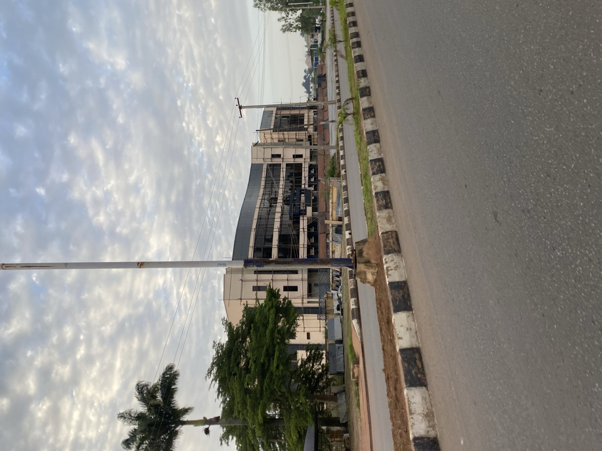 The image captures a street scene in Bar Jirgi Summa, Nigeria, with a modern building under construction in the background. The building features a contemporary design with large glass windows and scaffolding indicating ongoing work. To the left of the building, a tall palm tree stands alongside a lush green tree. In the foreground, a paved road runs alongside a black and white striped curb, with a brown, dirt embankment bordering the curb. Utility poles and wires are visible, suggesting the presence of infrastructure. The sky is partly cloudy, with a mix of white and gray clouds. The overall atmosphere is one of urban development and ongoing construction in a semi-tropical environment.