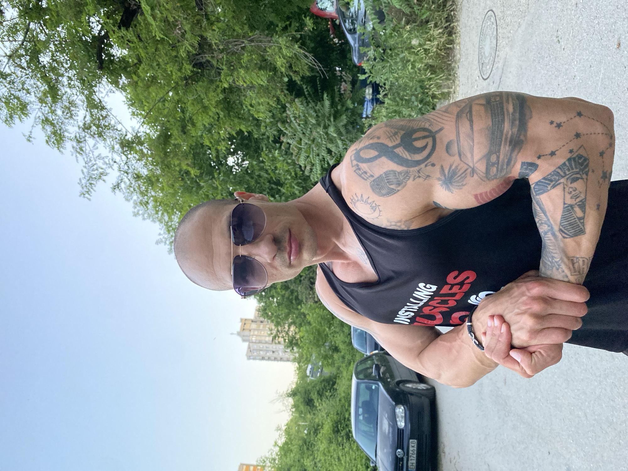 A bald, muscular man stands in an outdoor setting, likely Pleven, Bulgaria. He wears sunglasses and a black tank top. Tattoos cover his arms. He is standing on pavement next to a green area with trees. Cars are parked nearby. Tall buildings rise in the background. He clasps his hands together.