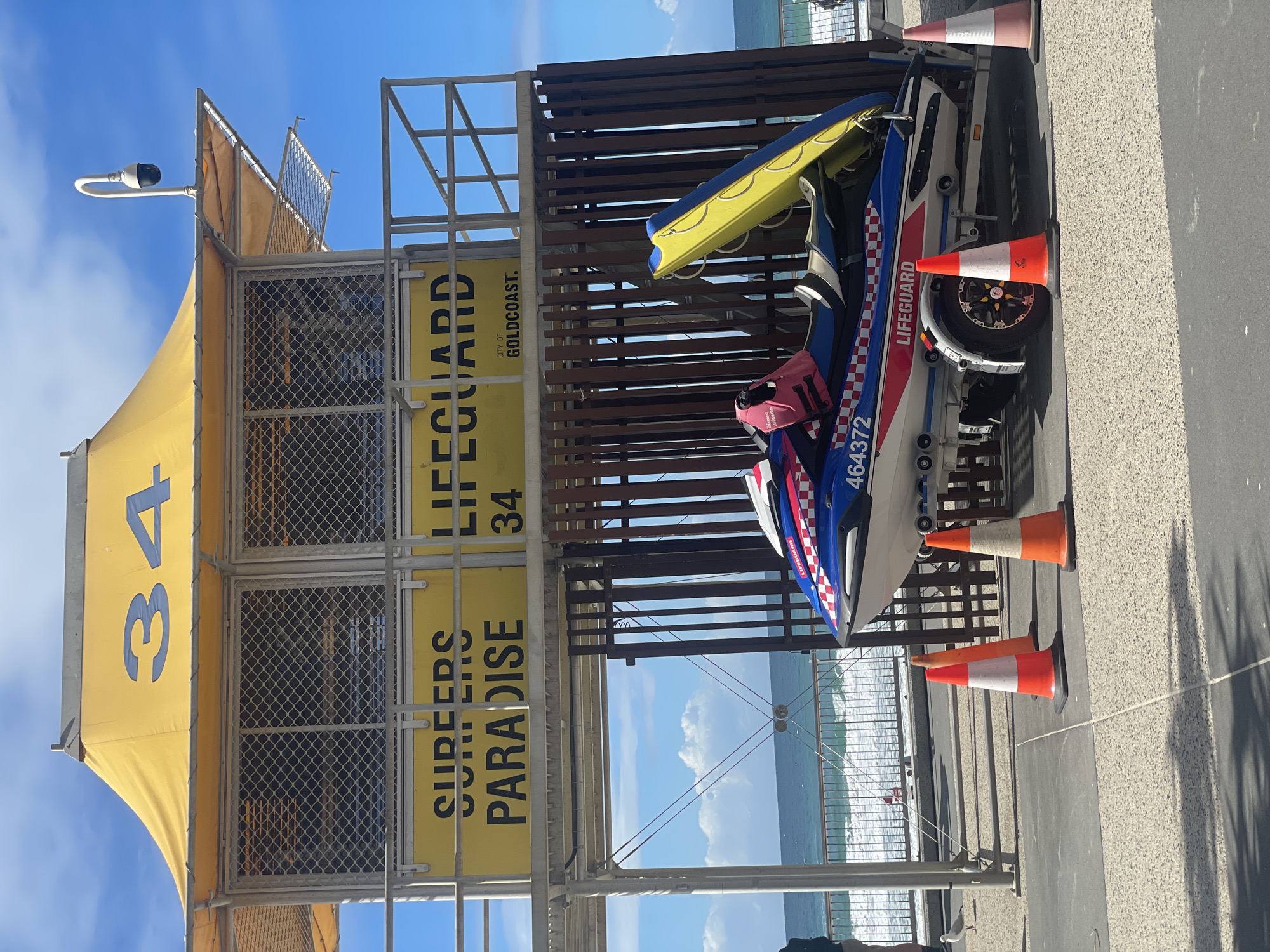 A bright yellow lifeguard tower numbered "34" stands against a blue sky with scattered clouds. The tower has "SURFERS PARADISE" and "CITY OF GOLDCOAST" signage. In front of the tower, a blue and red jet ski is parked on a trailer. The jet ski has "LIFEGUARD" and a number "464372" printed on its side. Orange and white traffic cones are placed on the ground around the jet ski. The background shows a sandy beach and the ocean with waves.