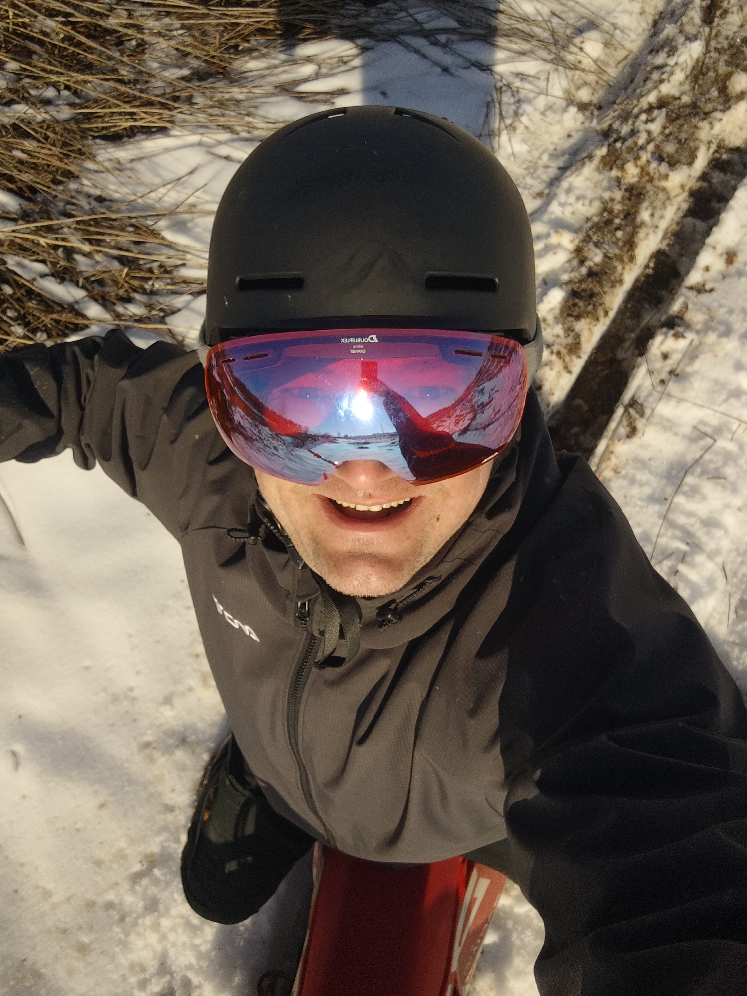 A man is taking a selfie while sitting in the snow. He is wearing a black helmet and red-tinted ski goggles, a black jacket, and black boots. He is sitting on a red sled. The reflection in his goggles shows a snowy landscape with trees or vegetation. The ground is covered in snow and dry vegetation. A tree branch is visible in the background. The photo was taken in the city of Прикамский, Russia.