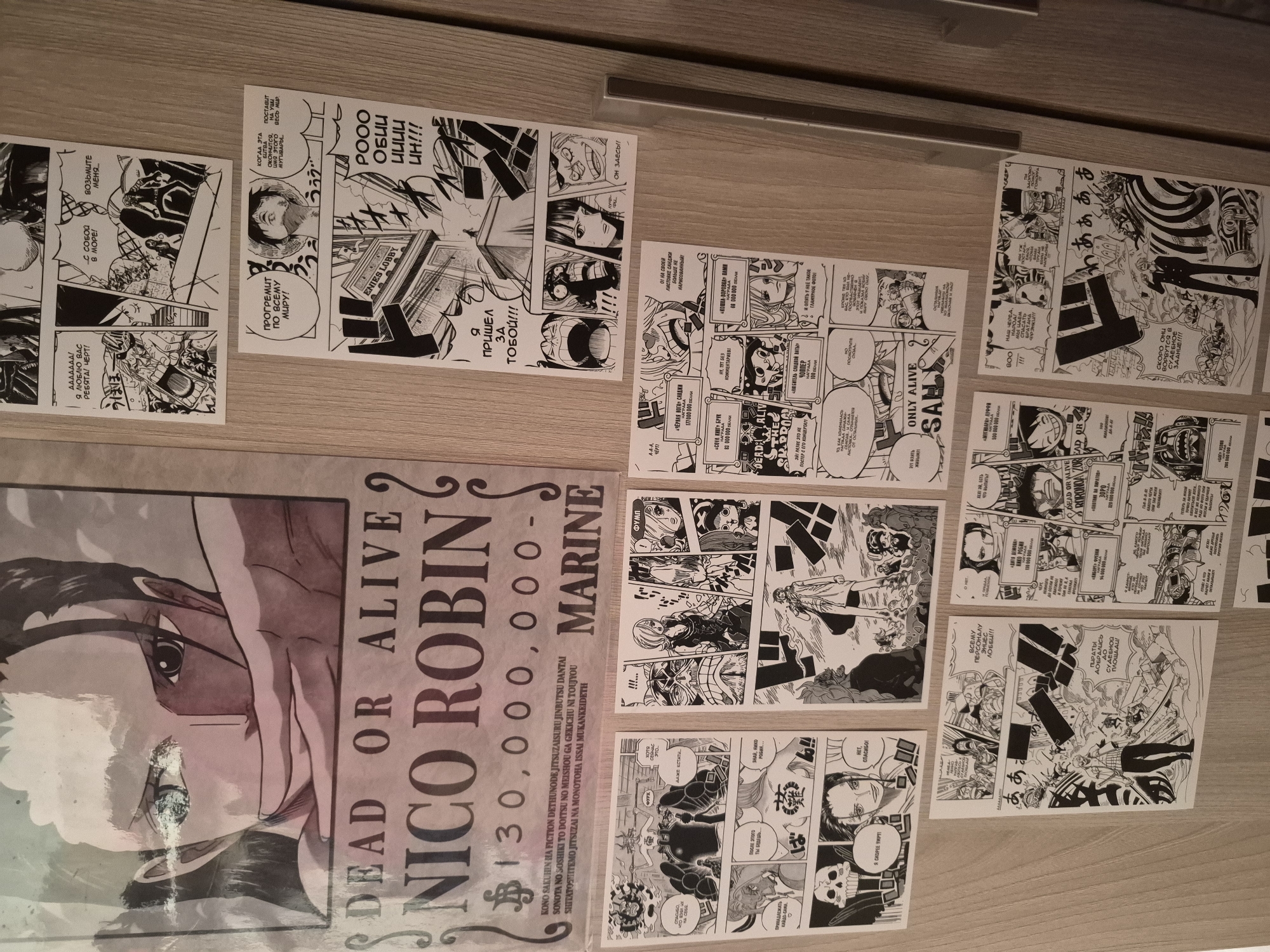 The image shows a collection of One Piece manga panels and a wanted poster for Nico Robin laid out on a light brown surface, likely a wooden table. The Nico Robin poster features her image in sepia tones, with the words "DEAD OR ALIVE" and a bounty of "130,000,000" visible. The manga panels are black and white, and appear to depict various scenes and characters from the series. Text is visible on most of the panels. The poster is partially covered with a clear plastic film.