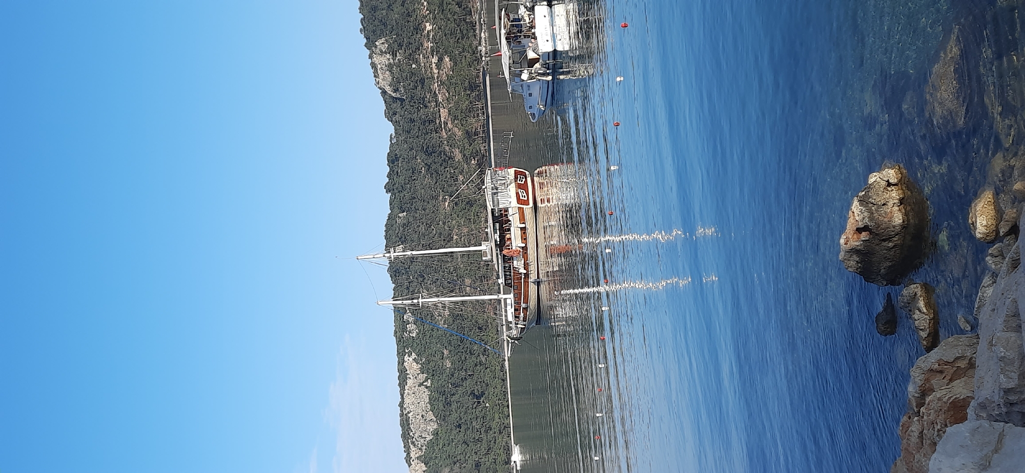 The image captures a daytime scene in Kemer, Turkey. A tall, brown, wooden sailboat with white masts is docked in calm, clear blue water, creating reflections. A forested mountain range is visible behind the boat. Several smaller white boats are docked further up the harbor. In the lower right corner, there are rocks along the shoreline. The sky is clear and blue.