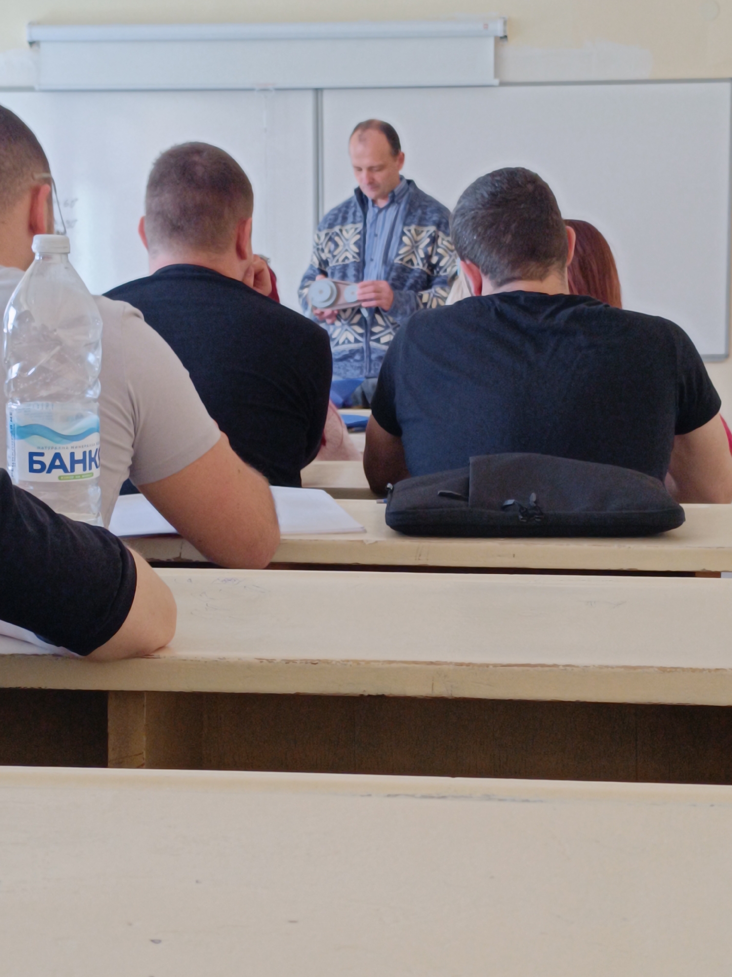 The image shows a classroom setting, possibly in an educational institution in Sofia, Bulgaria, judging by the "БАНК" water bottle visible. A man is standing in front of a whiteboard, holding a model that appears to be a mechanical component with belts and pulleys, suggesting a class in engineering or a related field. Several students are seated at desks, facing the front, their backs to the viewer. One student has a black zippered case on their desk. The desks are a light, worn color, and the overall lighting suggests an indoor environment. The water bottle on the left desk is branded with the "БАНК" logo, indicating it may be a local Bulgarian brand.