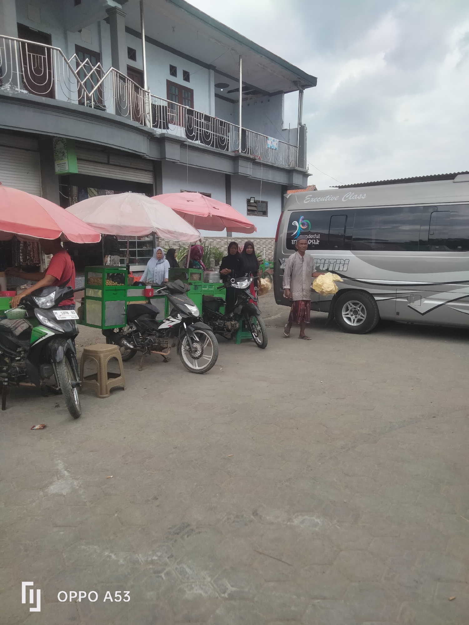 The image depicts a street scene in Badung, Indonesia, likely a market or roadside area. In the foreground, two motorcycles are parked. One is a green and black Honda motorcycle, and the other is a black and white motorcycle. Behind the motorcycles, several market stalls are set up under pink and white umbrellas. One stall displays fresh produce in green crates. Three individuals are visible near the stalls: two women wearing hijabs and a man in a light-colored shirt and patterned sarong. A large silver passenger van with "Executive Class" written on its side is parked on the right side of the image, obscuring part of the background. The architecture in the background suggests a multi-story building. The ground is paved with hexagonal stones. The sky is overcast.