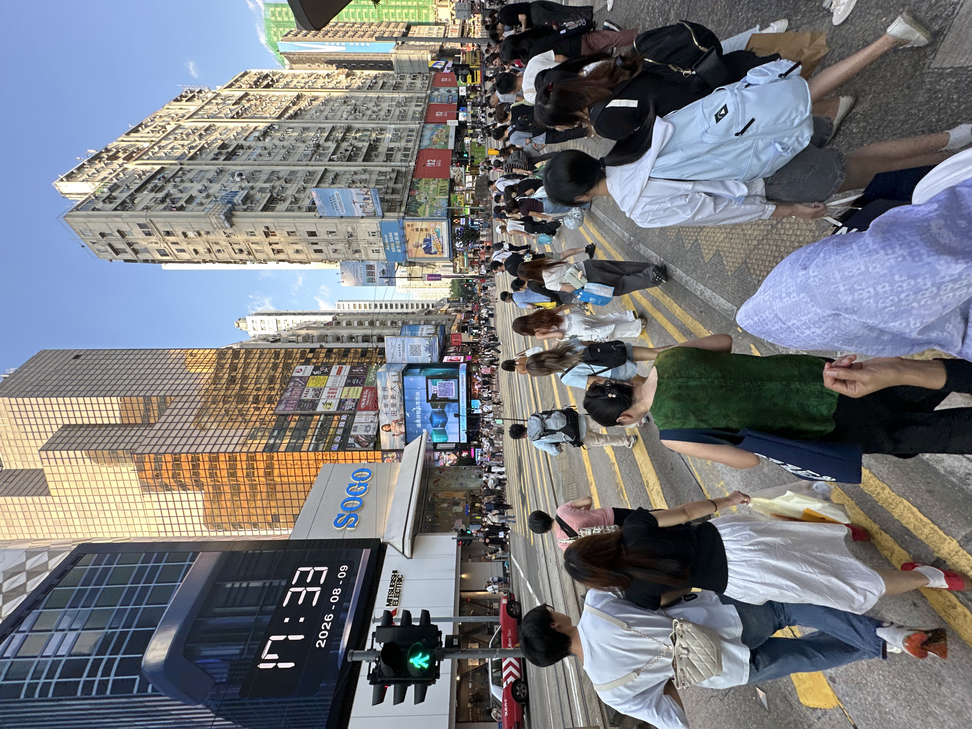 This is a street-level, medium shot photograph captured in Hong Kong, China, likely in the bustling Causeway Bay district given the presence of the SOGO department store. The image depicts a lively street scene with numerous pedestrians crossing a wide street marked with yellow and white pedestrian crossing lines. The foreground features a group of people walking away from the camera, engrossed in their movement across the crosswalk. A man in the immediate foreground wears a white t-shirt and blue jeans, carrying a light-colored bag over his shoulder. Next to him, a woman in a black top and white skirt walks with a shopping bag. Further back, a person in a green vest also carries a shopping bag.
The midground is filled with a crowd of people, suggesting a busy shopping area. In the background, the urban landscape is dominated by towering buildings. To the left, a prominent digital billboard displays the time "17:33" and the date "2026-08-09", alongside the SOGO department store logo. The buildings exhibit a mix of modern and older architectural styles, with many featuring glass facades reflecting the golden hues of the late afternoon sun. The sky is clear and blue. A red taxi and a traffic light with a green pedestrian signal are visible on the left side of the street. The overall atmosphere is one of urban dynamism and everyday life in a major city.
