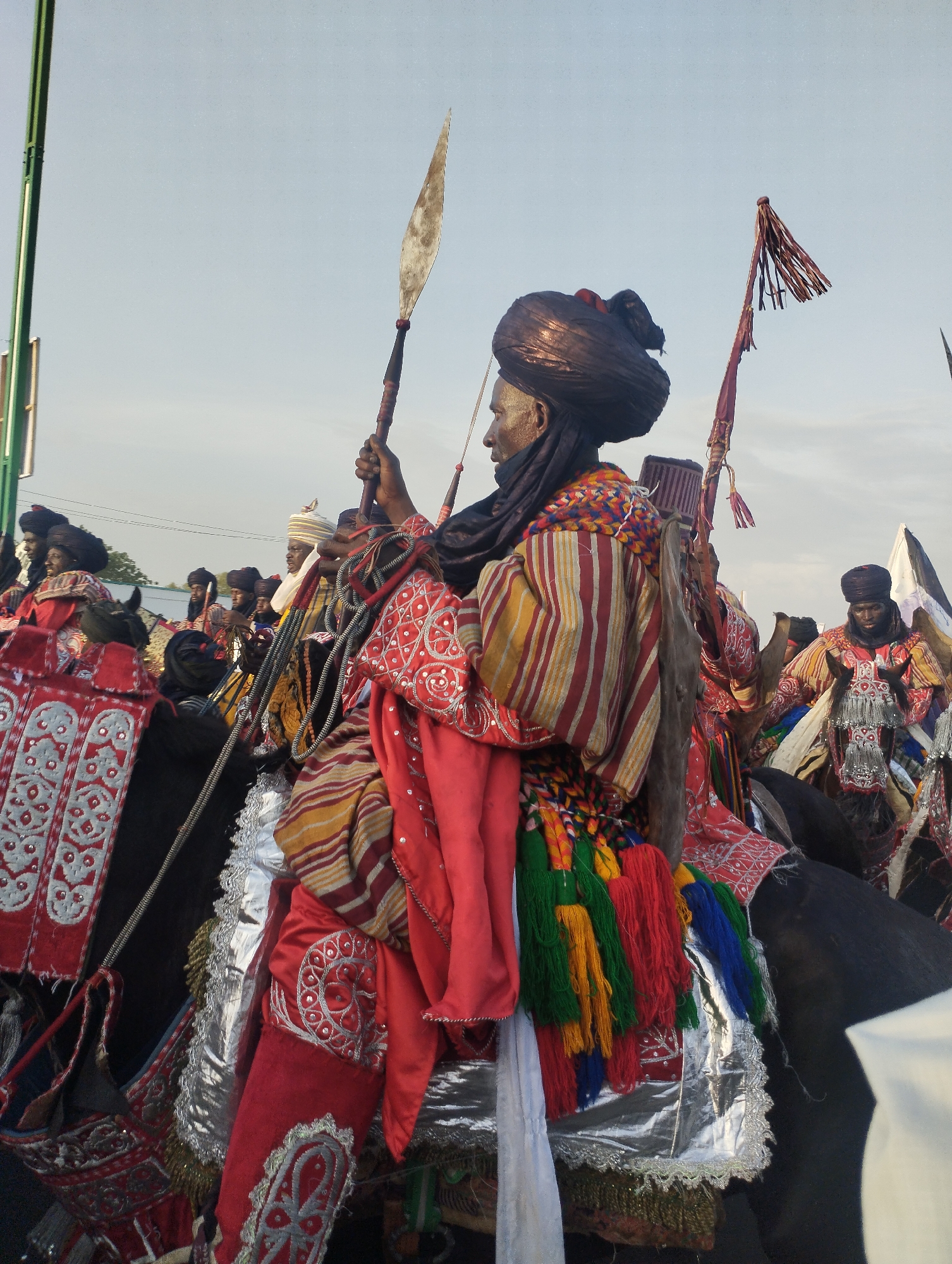 The image shows a group of men on horseback in Gombe, Nigeria, dressed in traditional attire. The central figure holds a spear, wearing a striped robe, turban, and colorful tassels. The horses are adorned with silver and red textiles. Other men on horseback are visible in the background, similarly dressed. The sky is overcast.