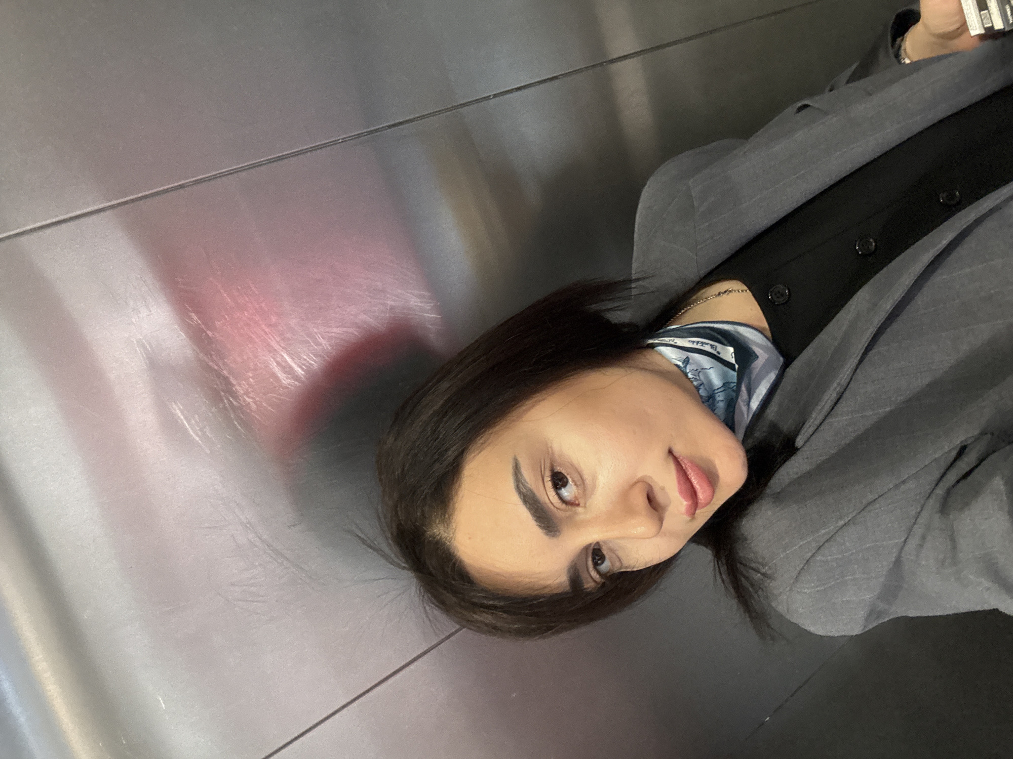 A woman with dark hair is wearing a grey pinstripe suit jacket, a black shirt, and a blue and white patterned scarf. She appears to be in an elevator with metal walls. The photograph may have been taken in Tbilisi, Georgia, though there is nothing in the image to confirm this.