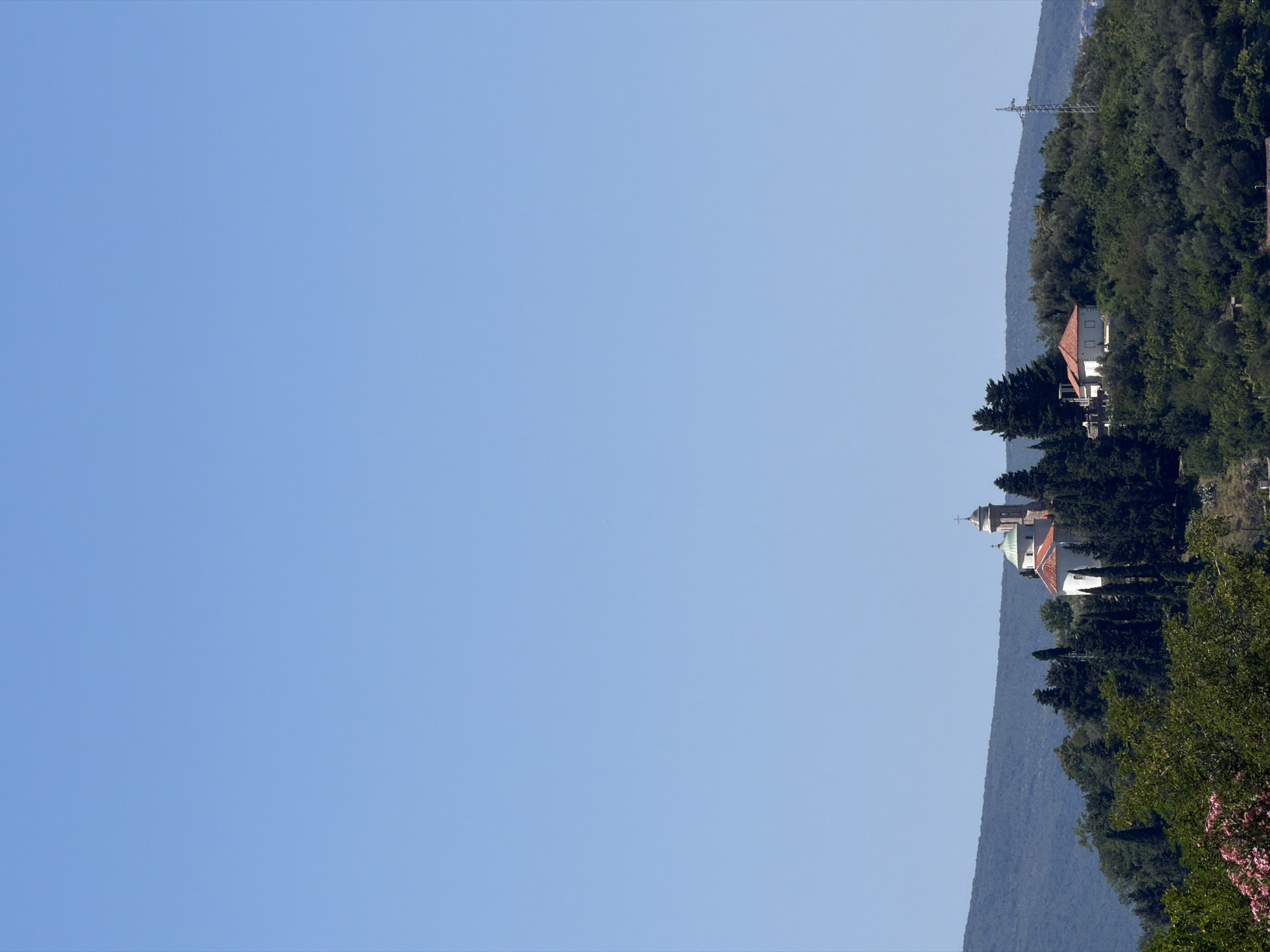 The image captures a serene view of the Church of St. Bartholomew in Lepetane, Montenegro, situated atop a verdant hill under a clear, pale blue sky. The church, a white structure with a red-tiled roof, is adorned with a dome and a bell tower topped with a cross. It is surrounded by a dense cluster of evergreen trees, including tall cypress trees that punctuate the landscape. In the foreground, lush green foliage and hints of pink flowers add a touch of natural beauty to the scene. To the right of the church, a smaller white building with a red roof is visible. In the distance, rolling hills covered in vegetation form a backdrop, and a solitary electricity pylon stands on the right horizon. The overall atmosphere is one of tranquility and natural beauty, highlighting the picturesque setting of the church.