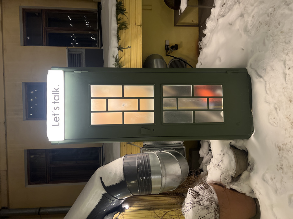 An olive-green booth-like structure stands with a sign that reads "Let's talk." above the door. The booth is situated outdoors, with snow piled around its base. To the right, a large accumulation of snow is visible. The background includes a yellow building with windows and a silver ventilation system partially covered with snow at the bottom.