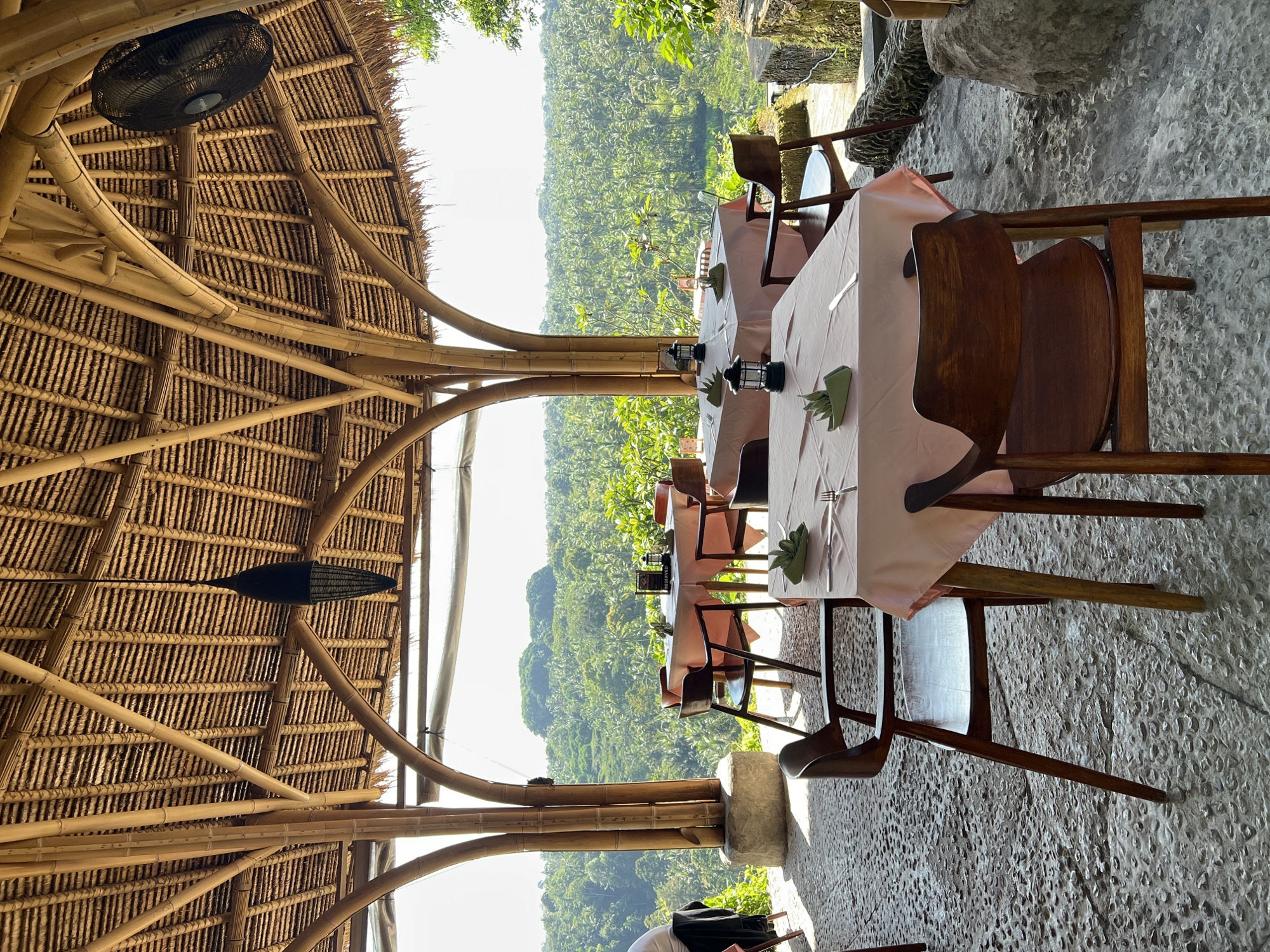 The image depicts a restaurant setting in Sayan, Indonesia, with tables set for dining under a thatched roof structure made of bamboo. The tables are covered with light pink tablecloths and feature place settings with silverware, napkins, and small lanterns. Brown wooden chairs surround the tables. The background showcases a lush, green landscape, typical of the region. A ceiling fan and a decorative ornament hang from the roof. The flooring is textured and made of stone. No people are prominently visible at the tables, suggesting a possible pre-service scene or a quiet moment between meals.