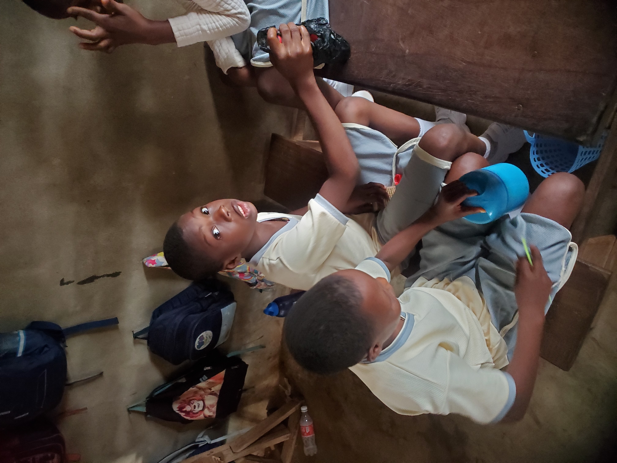 The photo depicts four school children sitting at desks in a classroom in Ukwu-Abwa, Nigeria. Three children are in school uniforms with gray shorts and off-white shirts. One child is turned towards the camera. Backpacks are on the floor near the children. A blue plastic basket, and a blue plastic cup are visible. A small bottle is on the floor.