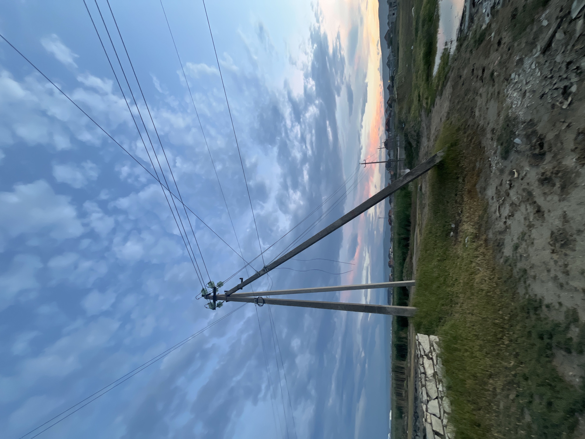 The image depicts a landscape with a prominent electric pole in the foreground. The sky is filled with clouds, some of which are reflecting the colors of the sunset. In the background, there is a residential area with houses. The ground is covered with grass and a dirt road. Based on the request, this image is taken in the city of Kaspiysk, Russia.