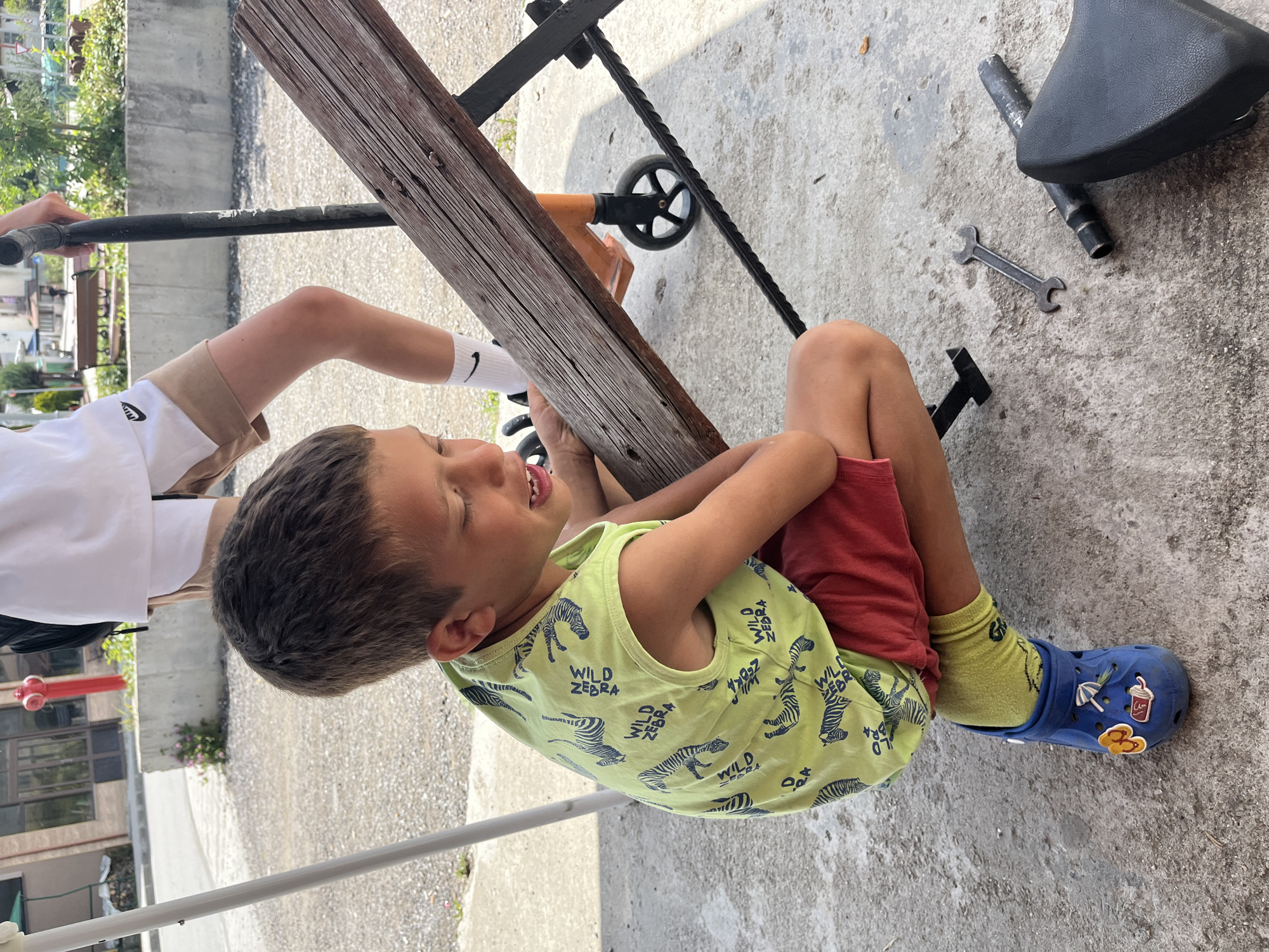 In the city of Barutin, Bulgaria, a young boy is crouched on a concrete surface, his eyes closed and mouth slightly open. He is wearing a green tank top with a zebra pattern, red shorts, yellow socks, and blue Crocs. To his right, a wooden plank rests on black metal supports, part of what appears to be a bench. Next to the plank, on the ground, lie a metal wrench and parts of a black seat, possibly from a bicycle or scooter. In the background, a person wearing a white t-shirt and light brown shorts stands with a scooter. The ground is covered in gravel and concrete, with some vegetation visible. The scene suggests a moment of rest or a break from activity, possibly involving repairs or maintenance.