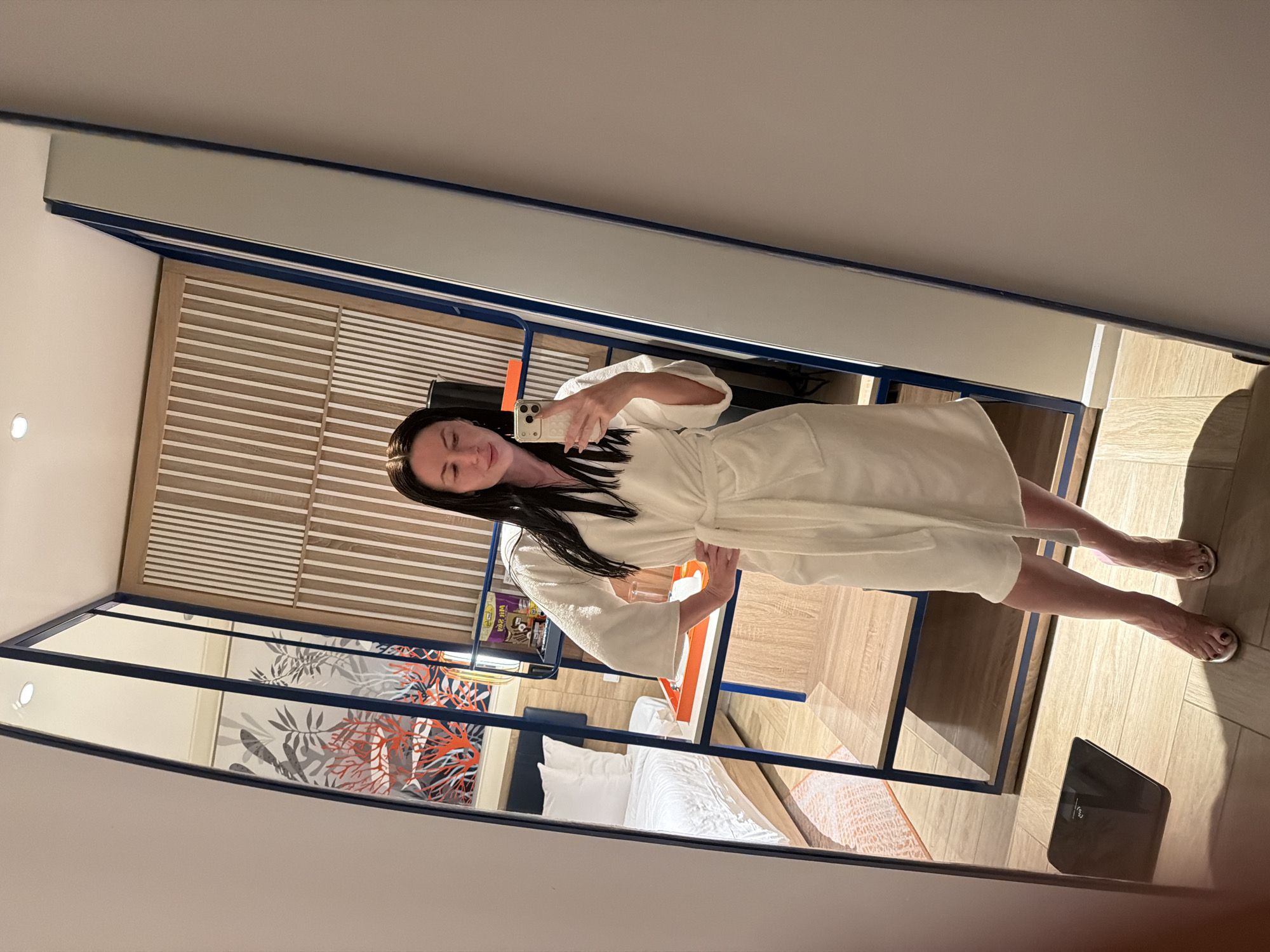 A long-haired woman, wearing a white hotel robe and clear high-heeled shoes, stands in front of a mirror, holding a white smartphone to take a selfie. Her left hand rests on her hip. The mirror reflects a modern hotel room interior. Visible in the reflection is a bed with white linens, a wall featuring an artistic motif of red coral and grey foliage, and a blue-framed shelving unit. On a shelf, an orange tray holds a glass, and a yellow "KOALA'S MARCH" package is present. Behind the shelving, a light wooden wall with vertical white stripes is visible. The floor is covered with light-toned, possibly wood-look, tiles. A black digital bathroom scale rests on the floor in front of the mirror. A recessed ceiling light is also reflected. This scene is situated within Dong Ninh Hoa Ward, Vietnam.