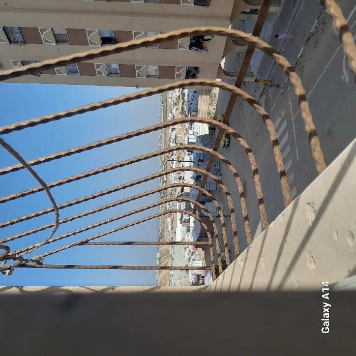 A view through a rusty, curved metal railing overlooking an urban scene. Several low-rise buildings and tents are visible in the distance under a clear blue sky. Adjacent to the railing is a building with multiple windows and laundry hanging. A road with pedestrian crosswalks and people walking is also in view. The foreground includes parts of the building's structure, casting shadows. The image also contains a "Galaxy A14" watermark on the lower right.