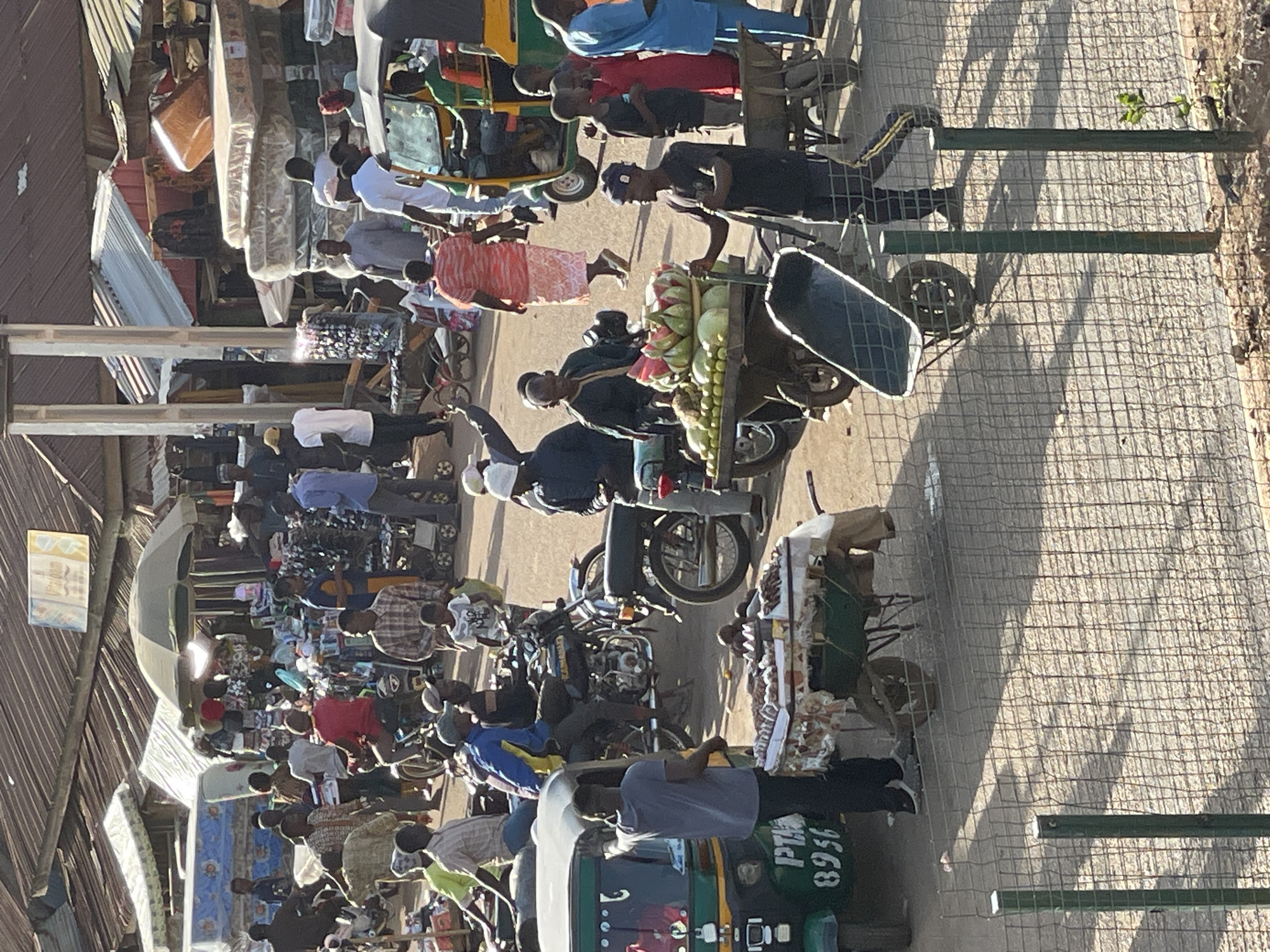 The image depicts a bustling street in Bukuru, Nigeria. A dense crowd of people is visible, many of whom appear to be engaged in buying or selling goods. Several motorcycles are parked or moving through the street, alongside a green auto-rickshaw. A vendor is transporting a cart laden with watermelons. Shops and structures with corrugated metal roofs line one side of the street. Sunlight is visible, suggesting a daytime scene.