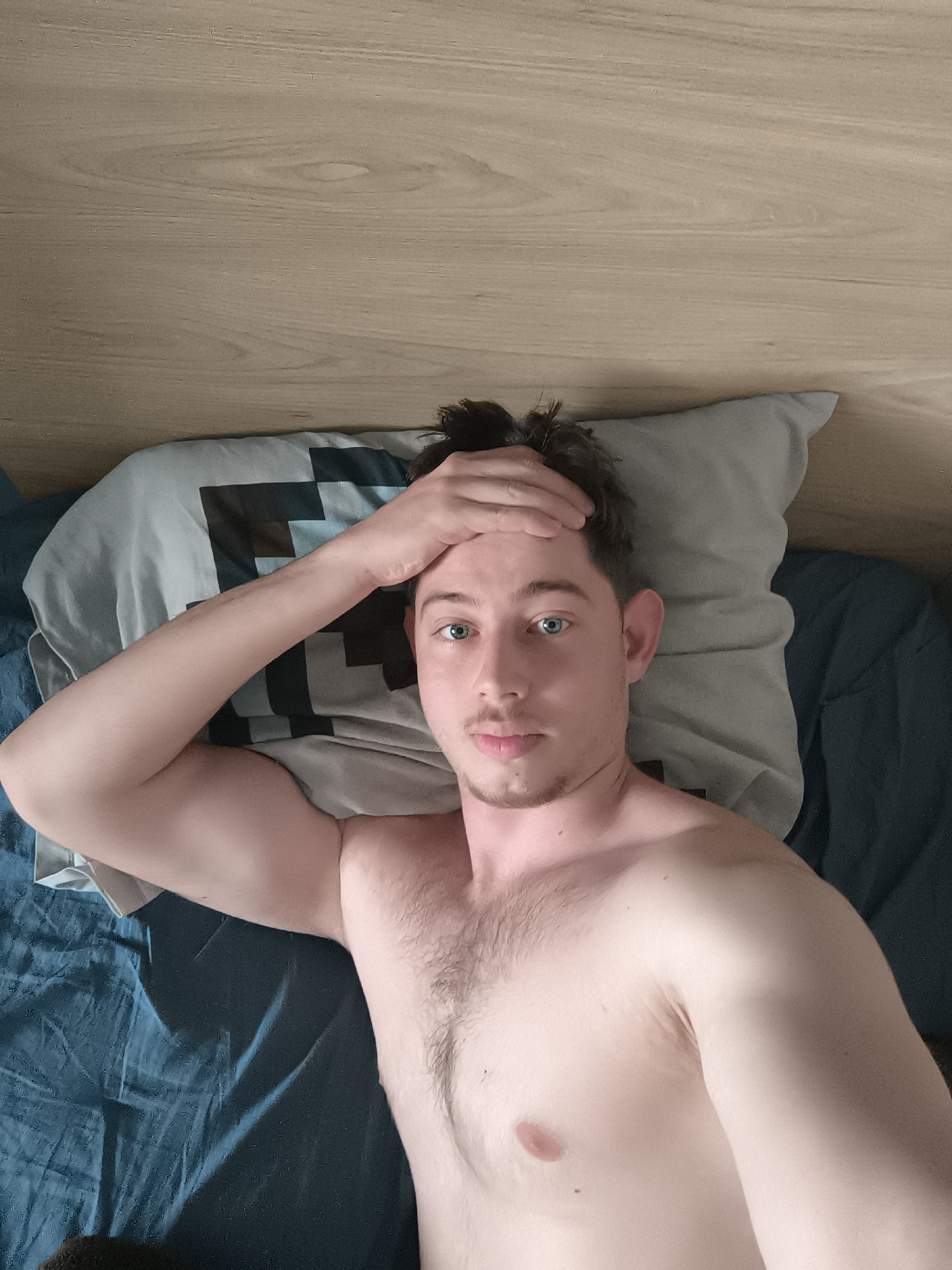 A shirtless man with fair skin, short brown hair, and blue eyes lies on a bed in what appears to be an interior setting. He rests his left hand on his forehead. He is on a pillow with a patterned design (likely black and grey squares) and the bed is covered in blue sheets. The headboard behind him is light-colored wood. The location is not discernible from the image. There is no indication of the location of Sofia, Bulgaria.