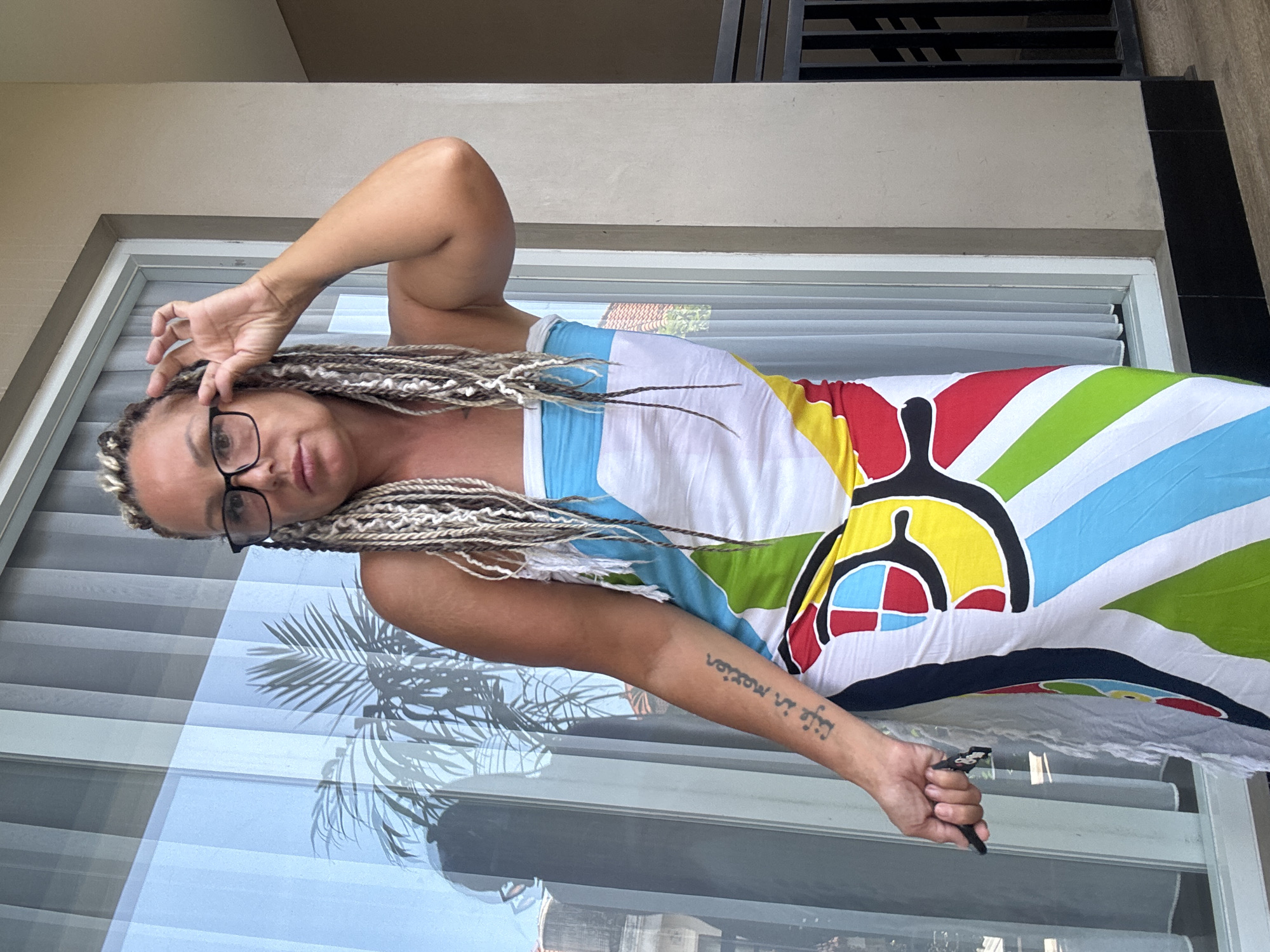 The image depicts a woman with blonde-streaked braids and glasses, wearing a colorful patterned dress, standing on a balcony. Her left hand is near her face, adjusting her glasses. Her right arm has a tattoo, and she is holding a black object (possibly a lighter or pen). The background shows window blinds, a palm tree reflection on the glass, and partial views of buildings. Given the location of Kuta, Indonesia, the setting is likely a hotel or residential building with a balcony overlooking an urban environment.
