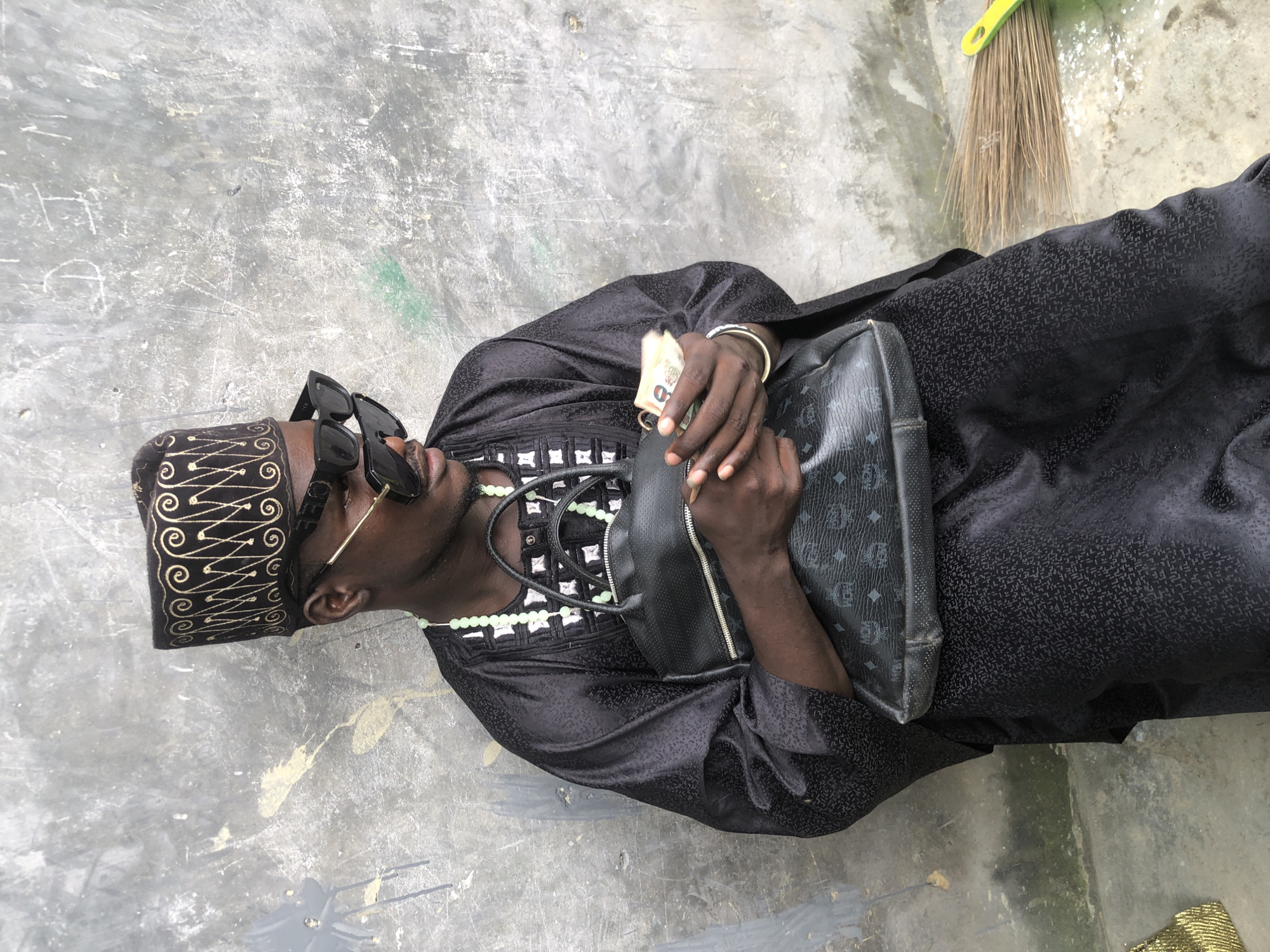 A man sits leaning against a textured grey wall in Ado, Nigeria. He wears a black, patterned traditional outfit and a black, embroidered cap. Oversized black sunglasses with gold accents are perched on his face. A necklace of light green beads is visible around his neck. He holds a black handbag with a distinctive MCM logo pattern and has a wad of cash in his hands. To the right, a broom lies on the ground. The wall behind him is weathered and shows signs of graffiti and discoloration.