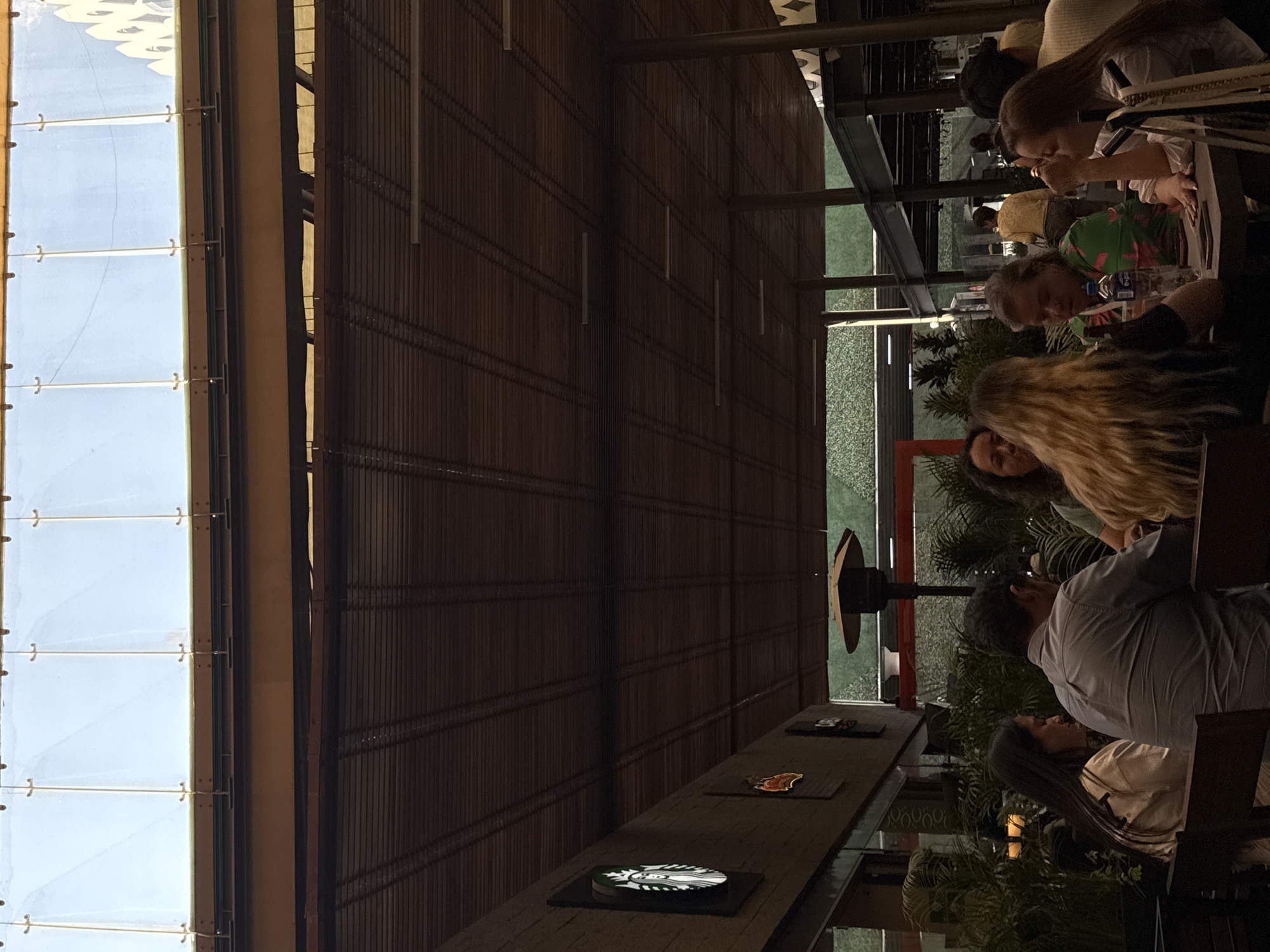 A casual outdoor scene depicts patrons at a Starbucks, likely in Puebla, Mexico. People are seated at tables, engaged in conversation and drinking. The environment has a bamboo-like wall structure, large windows showing the sky, and greenery adding ambiance. Starbucks brand emblems are visible on the tables.