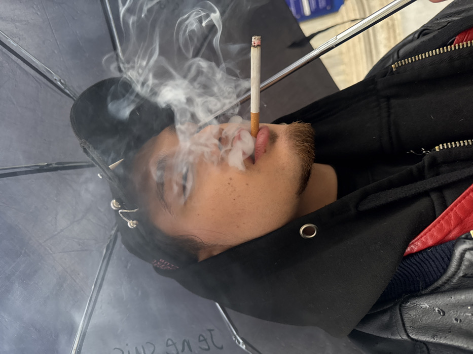 A young adult male is depicted in a close-up, chest-up shot, smoking a lit cigarette held between his lips and exhaling a plume of white smoke that partially obscures his face. He has light brown skin, dark hair, and a short goatee. He wears a black baseball cap adorned with multiple silver metal piercings and studs, including a spiral and a spike. Over a black t-shirt, he wears a black hooded sweatshirt with the hood pulled up, featuring a visible silver grommet. A black leather jacket, showing visible raindrops and a red panel or strap on the left side, is worn over the sweatshirt. The individual is positioned beneath a black umbrella, a segment of its metallic support rod visible on the right. Faint white French text, partially legible as "DU SAC," is visible on the underside of the umbrella's canopy on the left. The background outside the umbrella is indistinct, suggesting an outdoor environment, with a vague light-colored architectural element visible to the far right. The French text on the umbrella is consistent with a location in Paris, France, but a specific landmark or address cannot be identified from the visual evidence.
