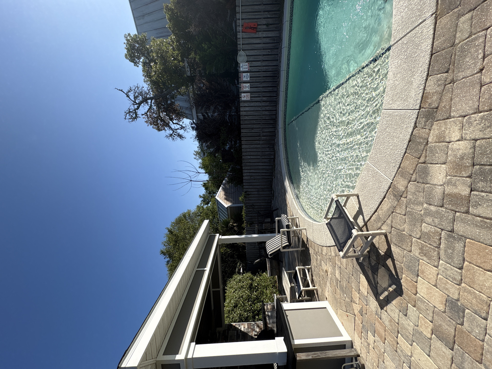 The image shows a section of a backyard in Panama City Beach, United States, featuring a swimming pool and patio area. The pool, filled with clear water, has a curved edge and is surrounded by a paved patio made of interlocking bricks. Several lounge chairs are positioned near the pool. Adjacent to the pool area is a wooden fence with pool safety signs. Beyond the fence is a mix of trees and vegetation. A white pergola structure stands on the opposite side of the patio, indicating a shaded seating or relaxation space. The sky is clear and blue, suggesting a sunny day. The overall scene presents a relaxed and private outdoor living space.