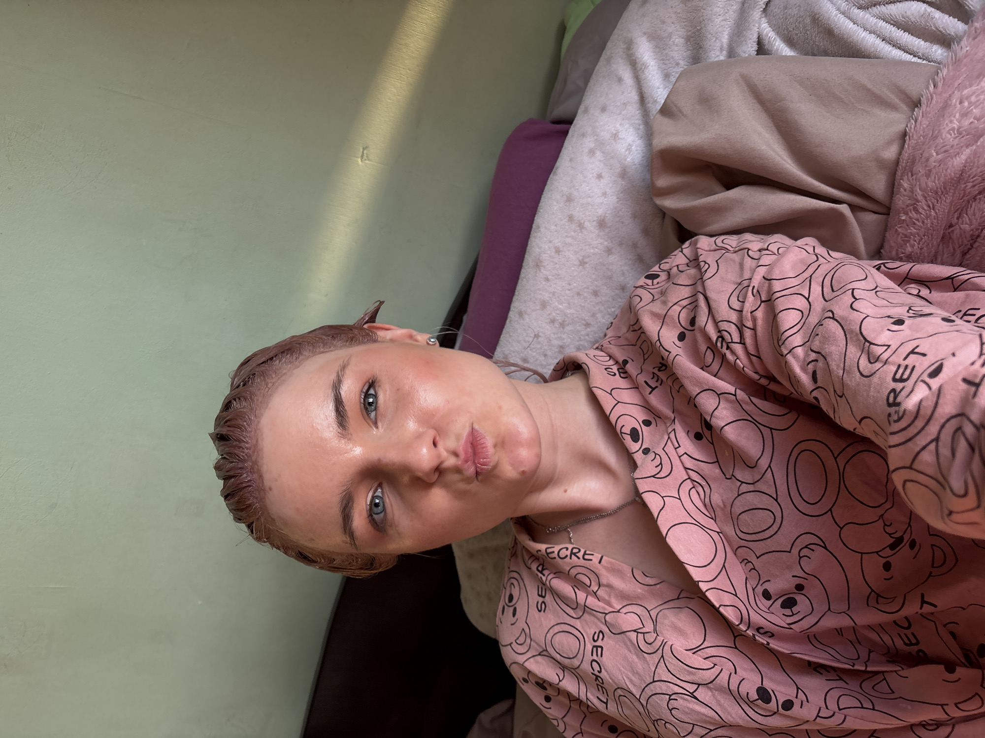 A young woman is depicted in an indoor setting, likely a bedroom, lying down and facing the camera with a playful pouty expression. Her light brown hair appears wet or treated, slicked back from her forehead. She has distinct blue eyes and her skin looks dewy. She is wearing a pink top or pajamas adorned with a repetitive pattern of black outline teddy bears and the word "SECRET." A silver chain necklace is visible around her neck.
The background features a light green wall, illuminated by a strong, diagonal beam of sunlight, suggesting either morning or late afternoon. Adjacent to her, an assortment of bedding is visible, including pillows and blankets in shades of beige, light grey (one with a star pattern), purple, and a fluffy pink texture. The overall impression is a casual, personal moment captured indoors in what could be a private residence in Ladozhskoe Ozero, Russia.