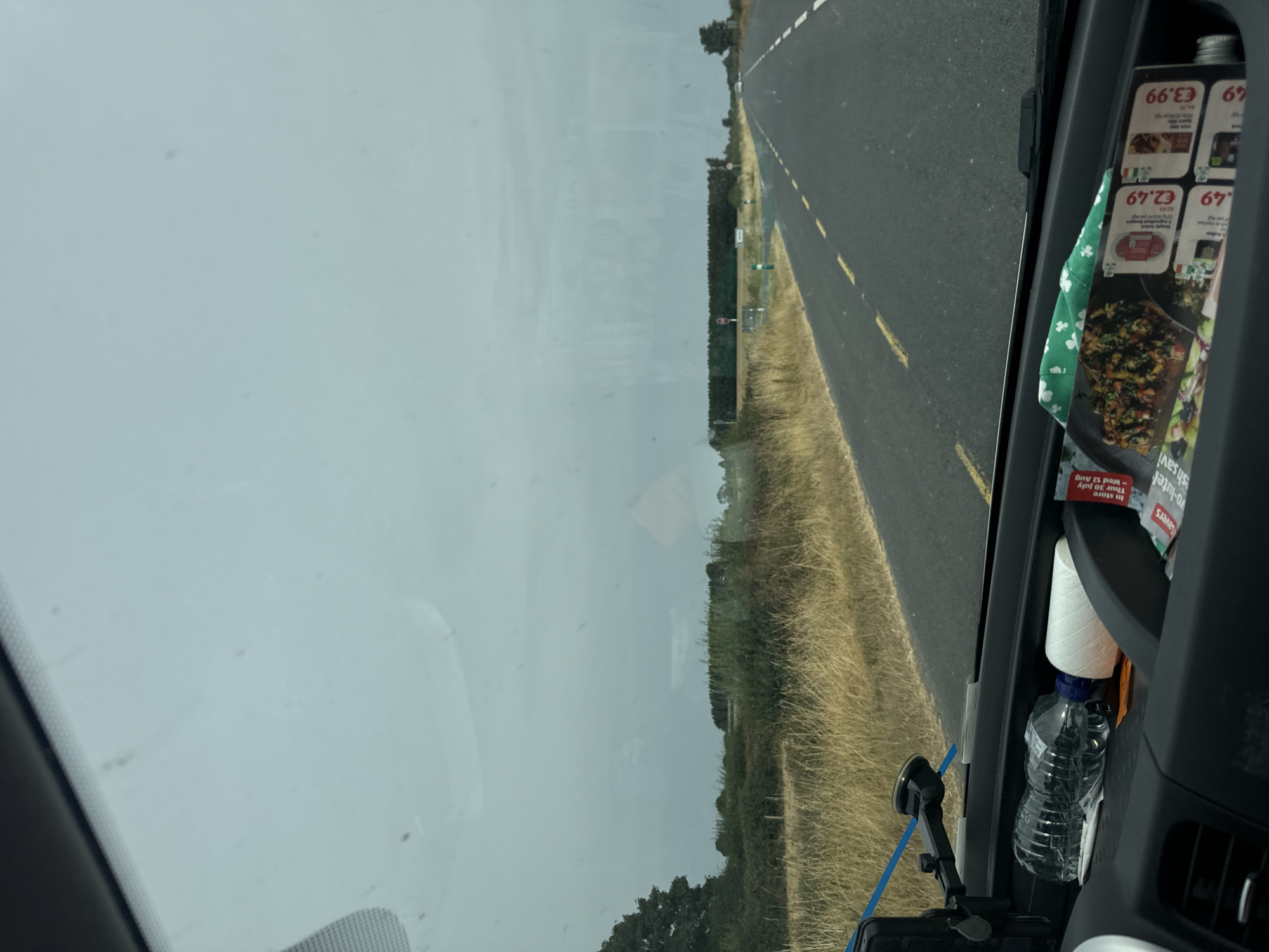 The image shows a view from inside a vehicle, looking out through the windshield. The sky is overcast and gray. In the distance, there is a flat landscape with trees and vegetation, and a road stretches into the distance. The road has a dashed yellow line marking the center. In the foreground, on the dashboard of the vehicle, there is a water bottle, a spool of white thread, and some brochures. One brochure has prices listed, including "€2.49" and "€3.99." The scene appears to be a rural road in the vicinity of Grevine, Ireland, given the presence of Irish prices on the brochure and the overall landscape.