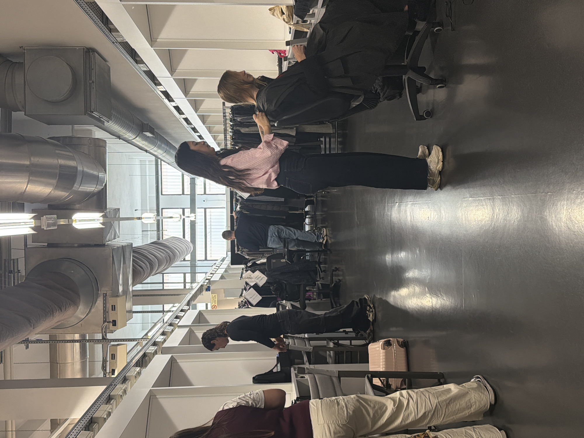 The photograph was taken indoors at a possible hair salon in Milan, Italy. The location appears to be a large, industrial-style space. The most prominent feature is the exposed ductwork and ceiling structure. Several individuals are present in the image. A woman with long dark hair, wearing a pink and white striped shirt and dark pants, appears to be styling the hair of a blond woman seated in a salon chair. A black cape covers the seated woman's attire. Further down the aisle, other people are seated, and some have luggage nearby. The floor is smooth and dark, reflecting the overhead lighting.