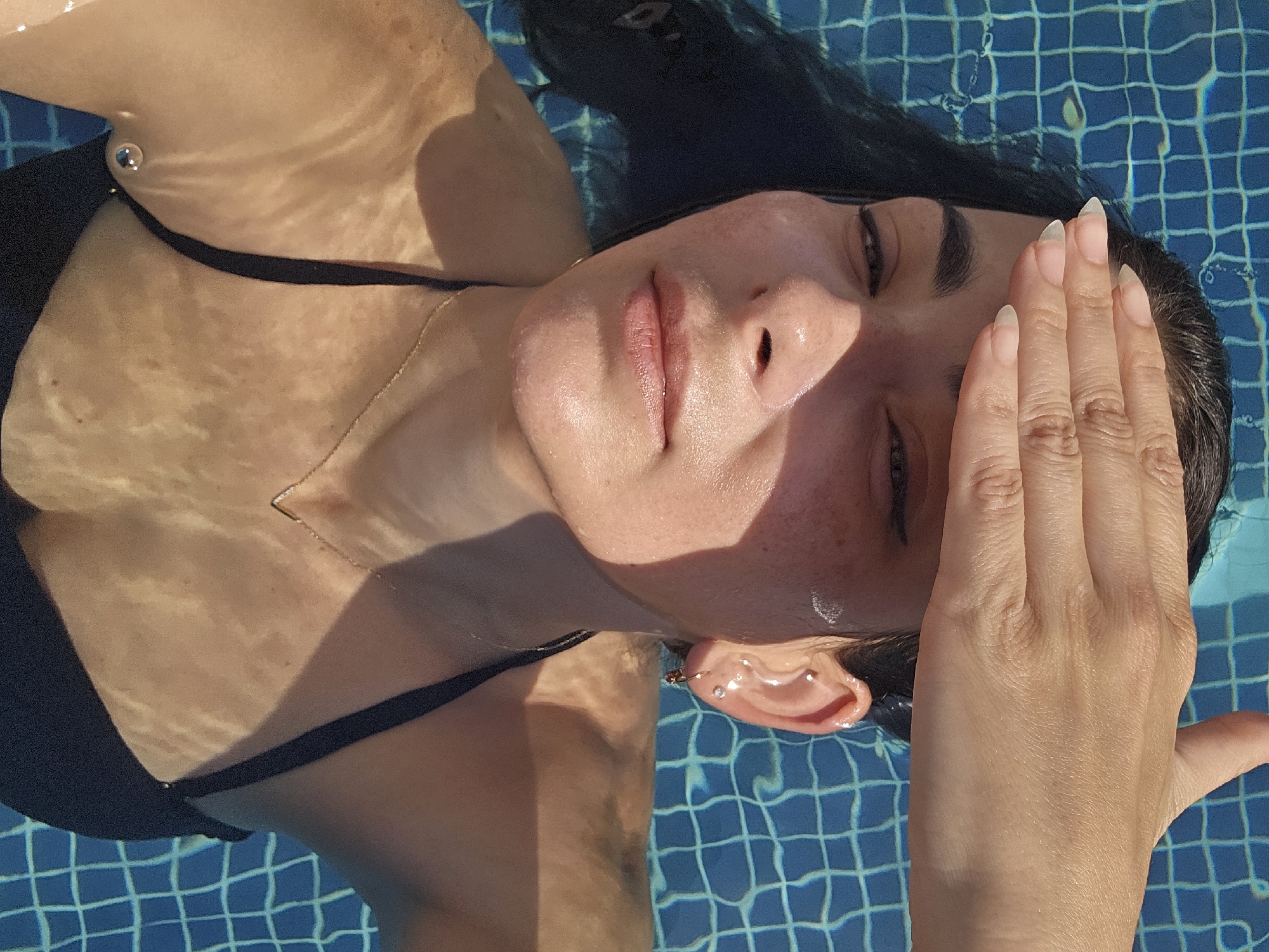 A woman with dark hair, wearing a black bikini, rests in a swimming pool. Her right hand is raised to shield her face from the sun. Sunlight reflects off the rippling water and onto her skin. The blue tiled bottom of the pool is visible beneath the water. The scene is set in Manama, Bahrain.