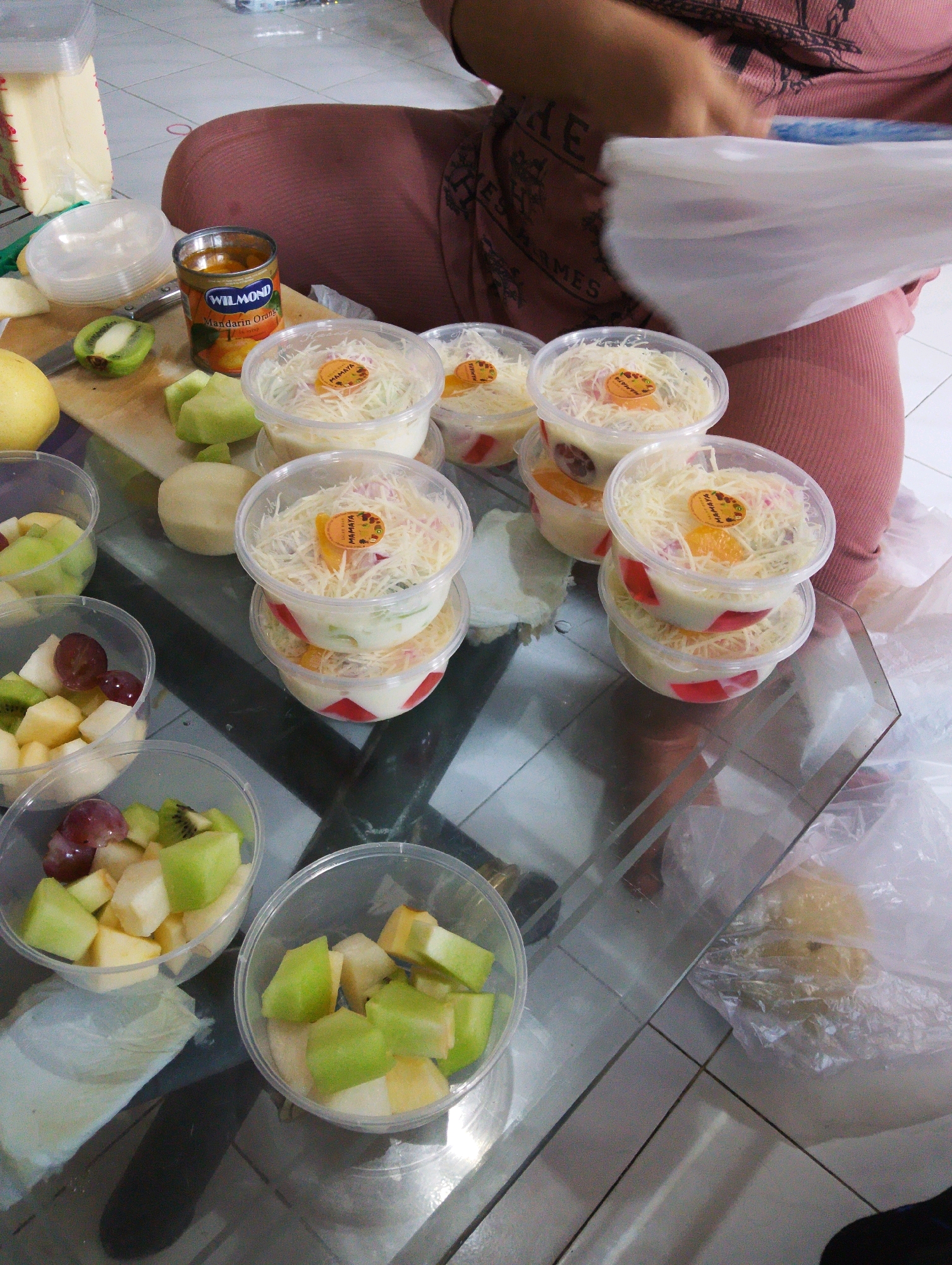 In Manaruwi, Indonesia, a close-up, slightly overhead shot captures a glass table laden with pre-portioned fruit salads in clear plastic containers. The fruits visible include chopped kiwi, melon, grapes, and possibly pears or apples, mixed with a creamy white dressing and topped with shredded cheese. Several containers are stacked, suggesting preparation for distribution or sale. A can of "WILMOND Mandarin Orange" is also present on the table, alongside a partially cut kiwi, a pear, and a block of cheese wrapped in plastic. A person, partially visible, is wearing a pink garment and is handling a white plastic bag, likely for packaging the fruit salads. The scene is set on a tiled floor.