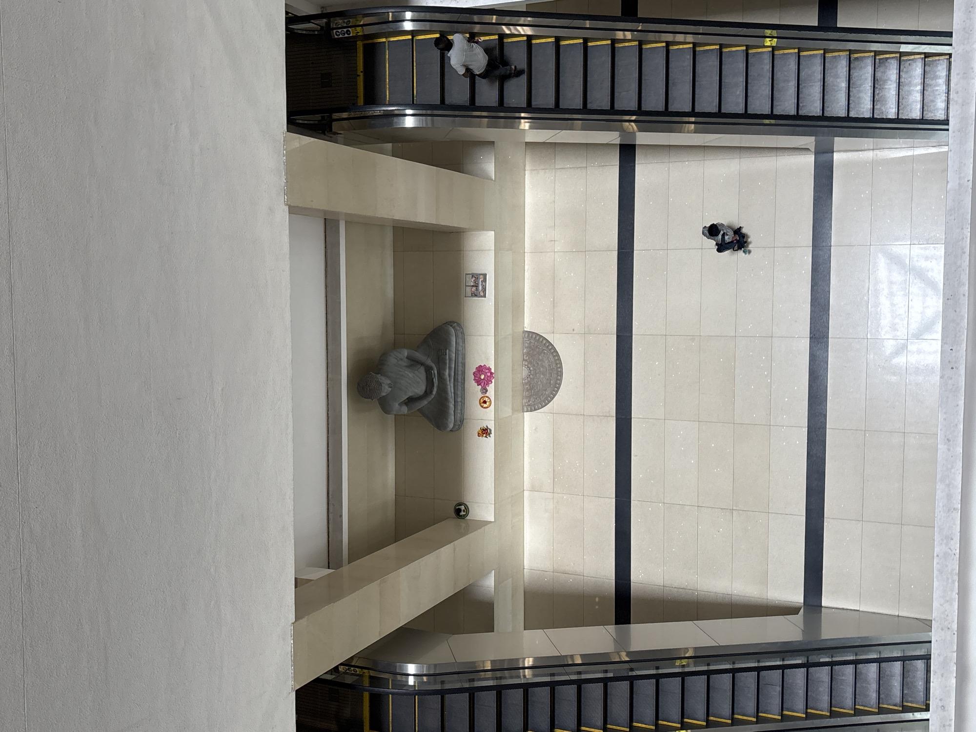 The image is an overhead shot within a building in Battaramulla, Sri Lanka. The scene includes an escalator on each end, both filled with black steps and bordered with yellow. There is a sculpture and what appears to be a design on the wall, in the middle. Two people are present; one on the top escalator and one in the main walkway. The walls and floors are mostly off-white.