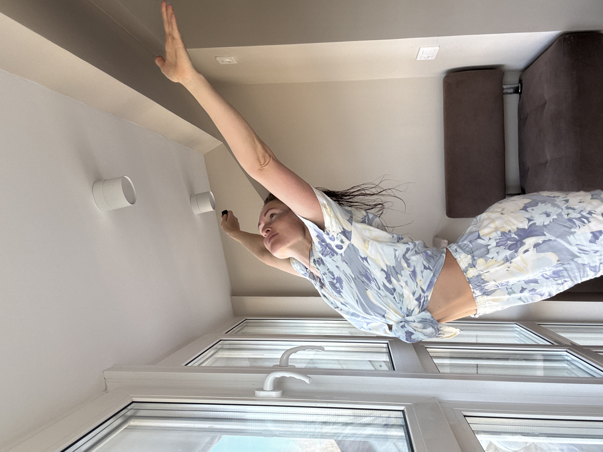 This media file captures a creative, rotated shot of a woman seemingly suspended in a room. The image is oriented vertically, making the woman appear to be "flying" or floating along the ceiling.
The main subject is a young woman, seen from the chest up, with her body angled. Her right arm is extended straight upwards, palm open, as if reaching for the ceiling, which in the rotated view becomes the upper wall. Her left arm is bent, and she holds a small, dark cylindrical object, possibly a remote control or a small flashlight. Her head is tilted back, and she looks upwards with a serene or focused expression. Her dark hair is fanned out around her head, enhancing the illusion of movement or being weightless. She is wearing light-colored, patterned pajamas or lounge wear; the top is tied in a knot at her waist, and the bottoms feature a blue, white, and yellow floral design.
The setting is an indoor room, likely a modern apartment in Chaikovsky, Russia. A large window with white frames and handles occupies the lower part of the image, suggesting bright daylight illuminating the scene from outside. The room features white walls and a ceiling with a brown-beige colored soffit or trim running along it. Two white cylindrical objects, possibly recessed lights or ventilation vents, are visible on the white wall. In the background, part of a brown upholstered sofa or armchair is visible, providing a hint of furniture in the room. The overall impression is one of playful levity, utilizing the unusual camera angle to create an artistic or whimsical effect. There is no visible text in the image.