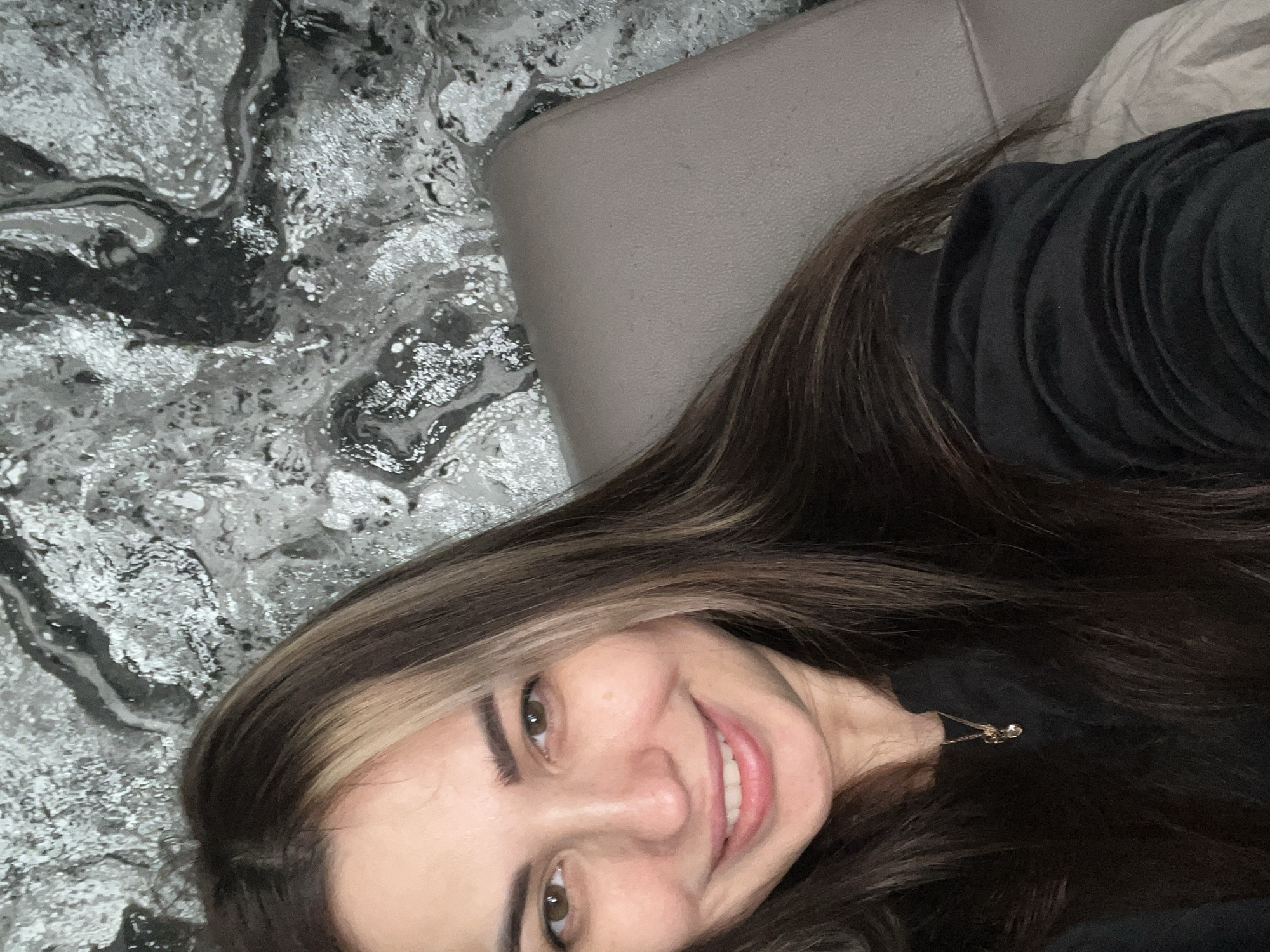A person is looking at the camera and smiling. They have long brown hair with blonde highlights. They are wearing a black shirt. The background includes a gray textured wall and a gray headrest. The location is Unknown, Unknown.