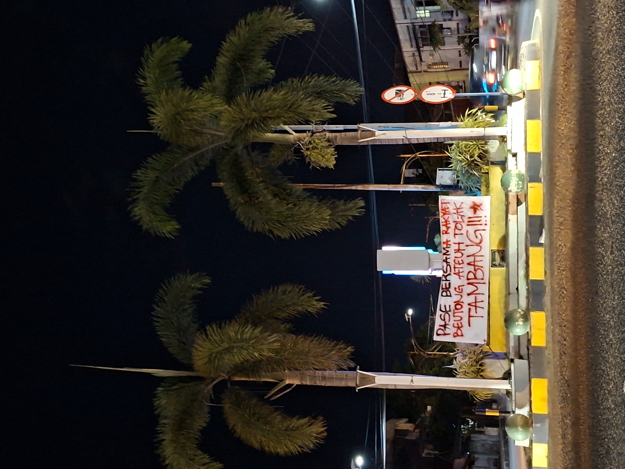 The image shows a street scene at night in Lhokseumawe, Indonesia. Two palm trees are visible on the left. A white banner with red text that reads "PASE BERSAMA RAKYAT BEUTONG ATEUH TOLAK TAMBANG!!!" is attached to a traffic barrier on the right. The traffic barrier is painted with yellow and black stripes. A road sign is visible behind the banner. A road and buildings are in the background. Cars are driving on the road. Streetlights illuminate the scene.