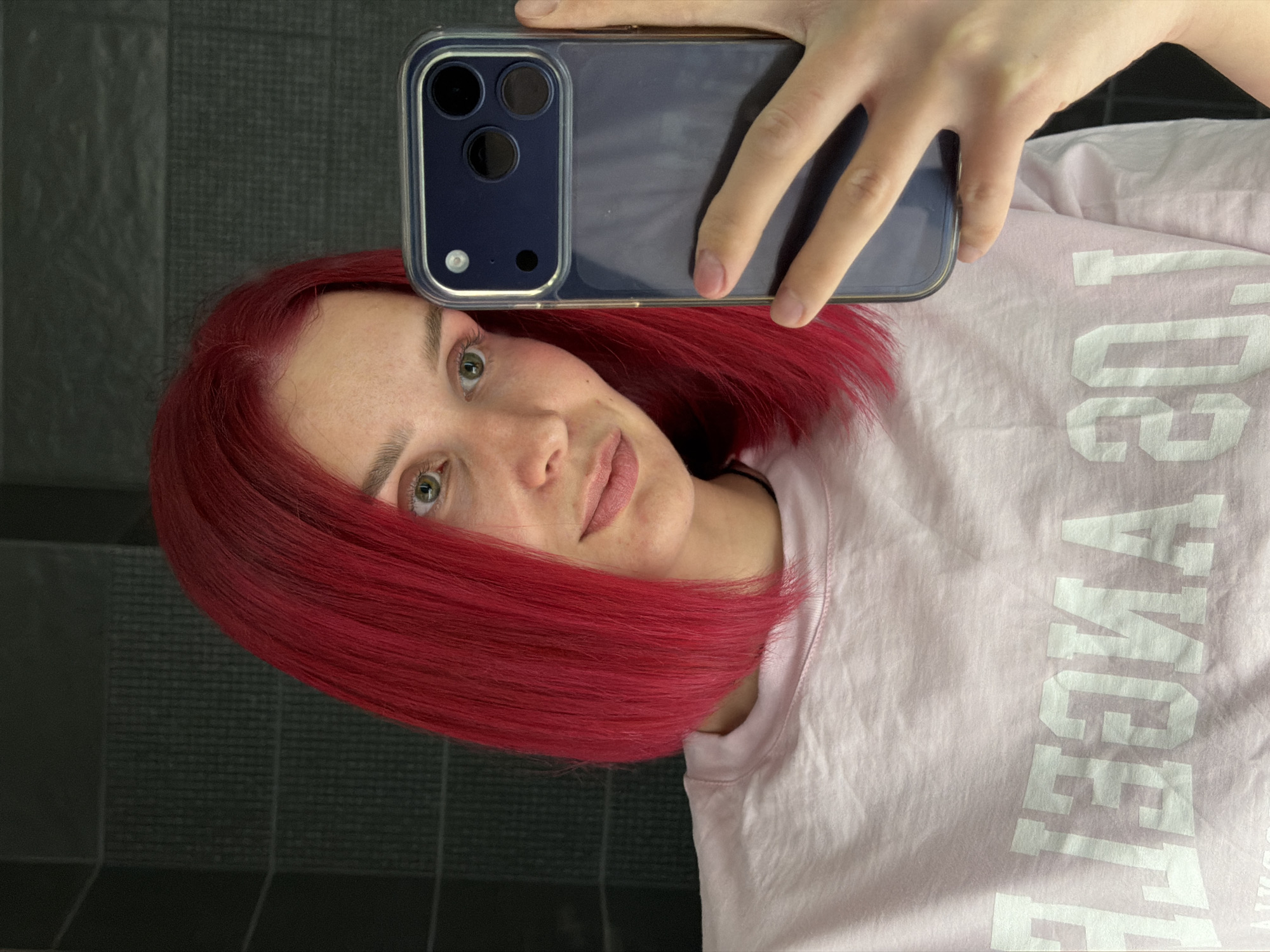 A woman with red hair takes a selfie using a smartphone. The phone, a dark blue model with a clear case, partially obscures her forehead. She is wearing a light pink t-shirt with the word "LEEGOMA" printed in white. The background shows dark grey, possibly tiled walls. It is not possible to determine the location in Chaikovsky, Russia, or any further details about the environment or the subject's actions or interactions from the image.